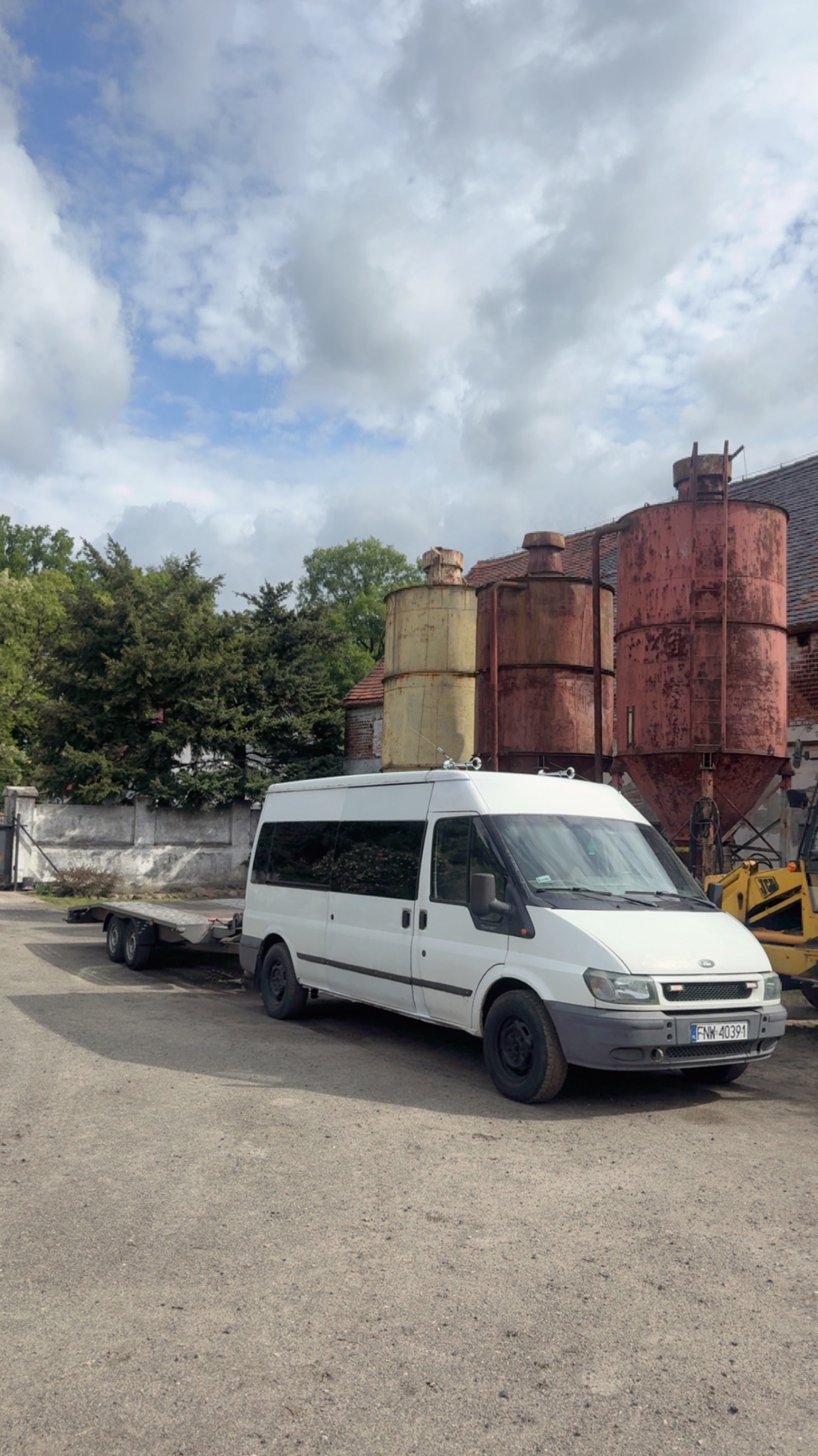 Biały bus z przyczepą lawetą na żwirowym placu, w tle silosy i budynek z cegły, częściowo widoczna koparka. Pochmurne niebo.