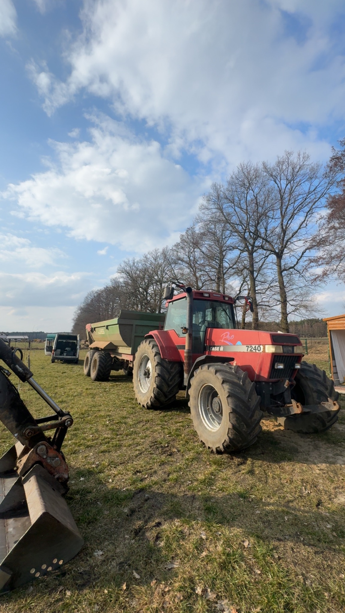 Czerwony traktor Case 7240 z przyczepą na łące, obok widoczna koparka. Błękitne niebo z chmurami w tle. Sprzęt gotowy do pracy.