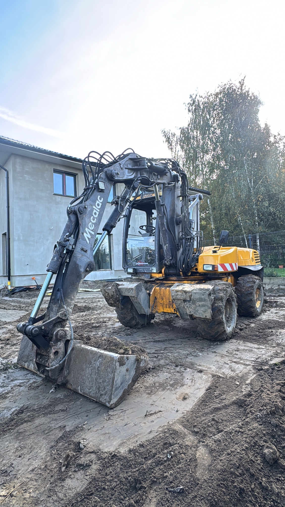 Żółto-szara koparka Mecalac na placu budowy, z widocznym budynkiem w tle i śladami opon w błocie. Maszyna gotowa do pracy.
