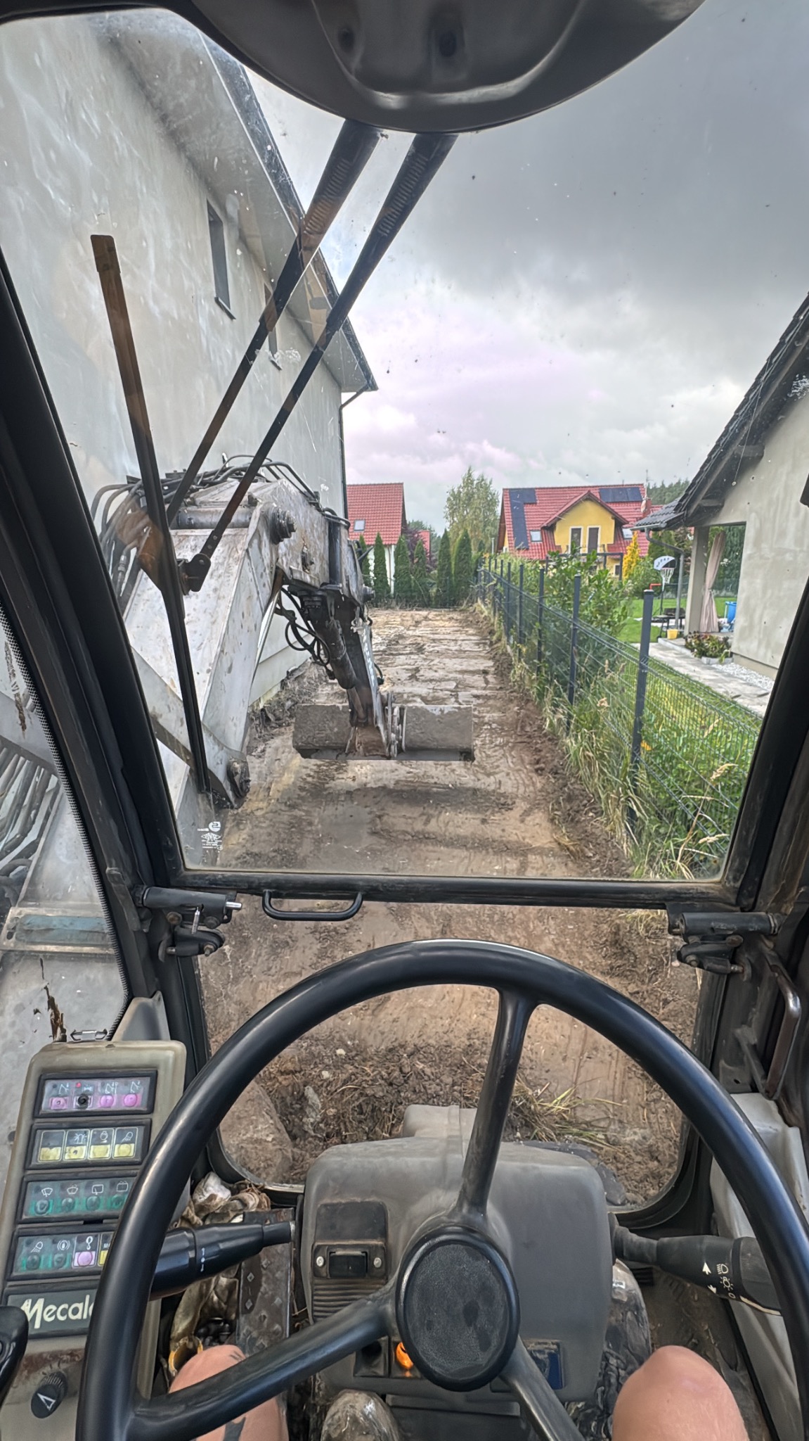 Widok z kabiny koparki podczas prac ziemnych na działce, zarys fundamentów, domy w tle. Perspektywa operatora, pochmurne niebo.
