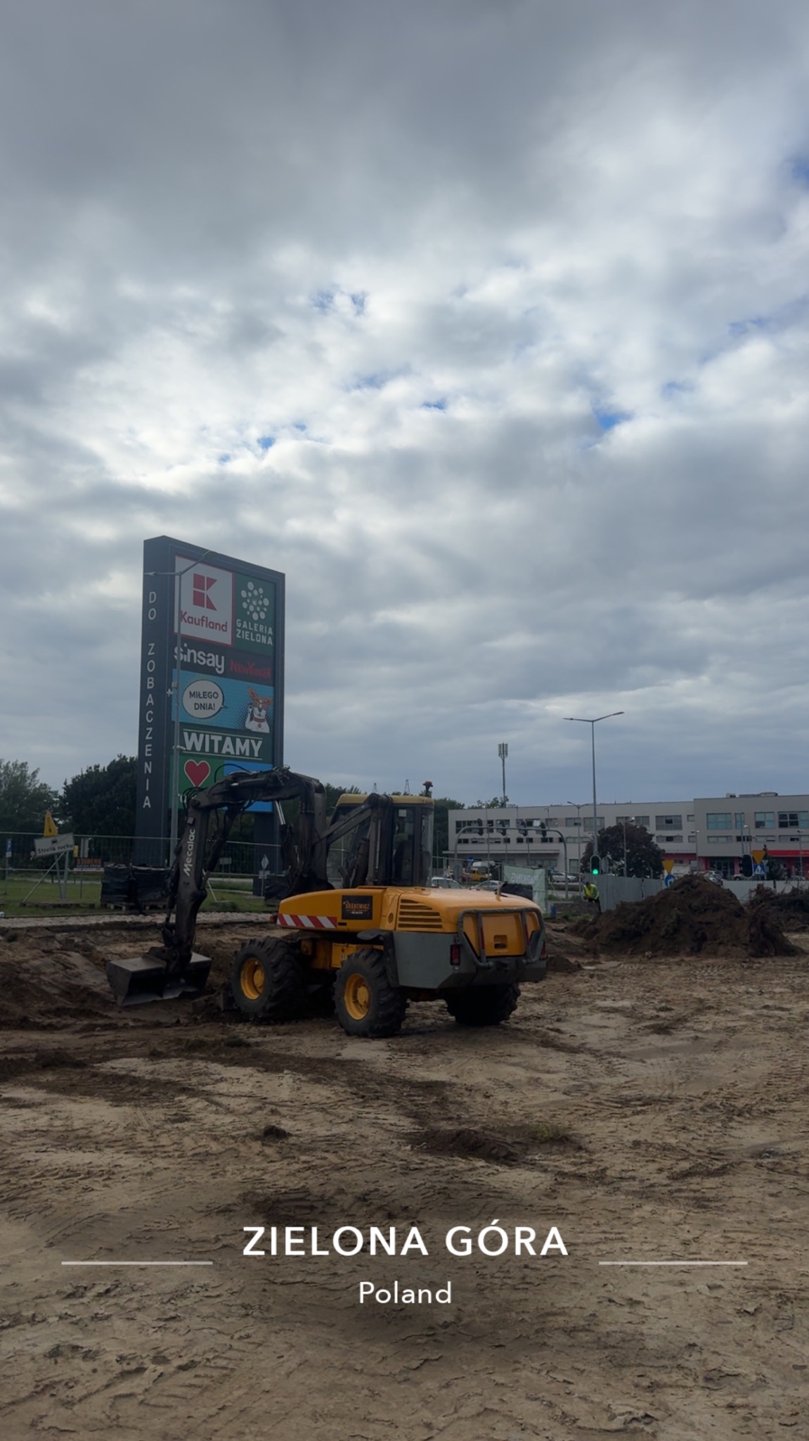 Żółta koparka Mecalac na placu budowy w Zielonej Górze, w tle tablica reklamowa galerii handlowej. Widoczne ślady pracy maszyny w ziemi.