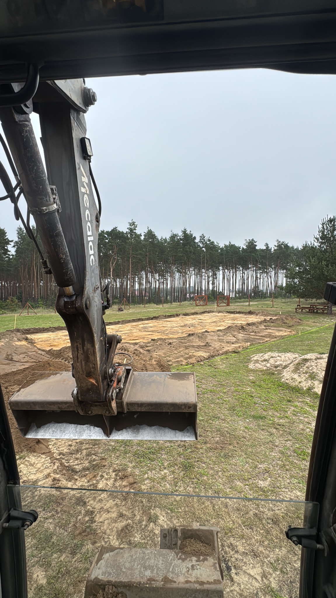 Widok z kabiny koparki na przygotowany teren pod budowę, z widocznymi wykopami i lasem w tle, w pochmurny dzień. Ramię koparki z łyżką w kadrze.