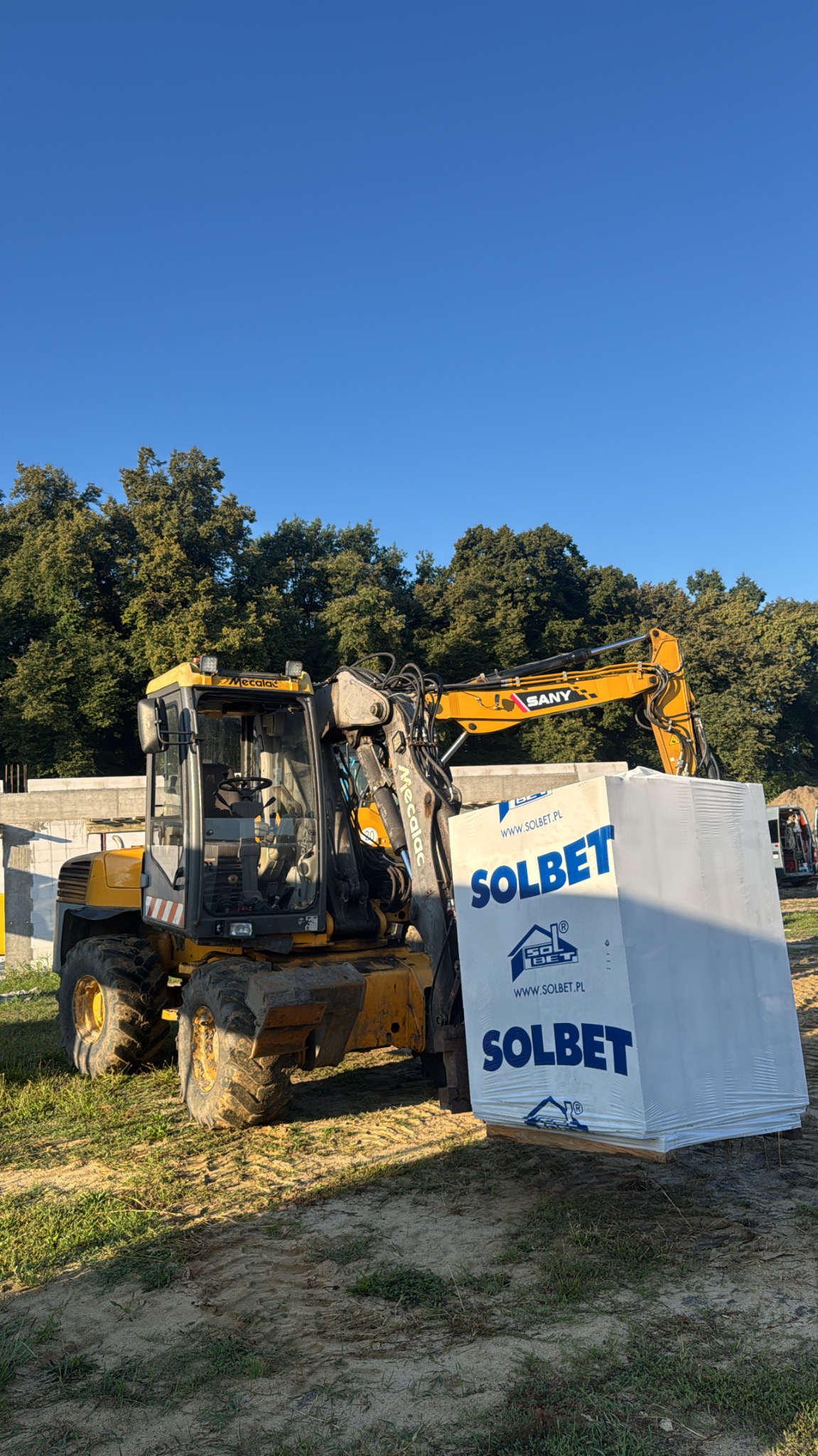 Żółta koparko-ładowarka Mecalac SANY przewozi paletę z bloczkami Solbet na placu budowy. W tle zieleń drzew i fragmenty betonowych fundamentów.