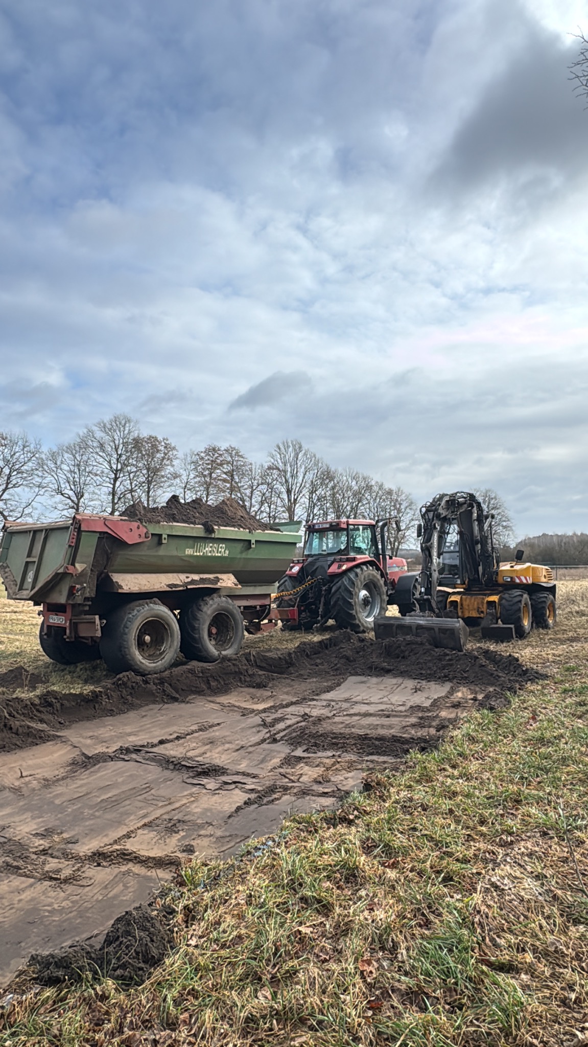 Czerwony traktor z przyczepą wypełnioną ziemią i żółta koparka na polu, ślady opon na ziemi, pochmurne niebo w tle. Prace ziemne w toku.