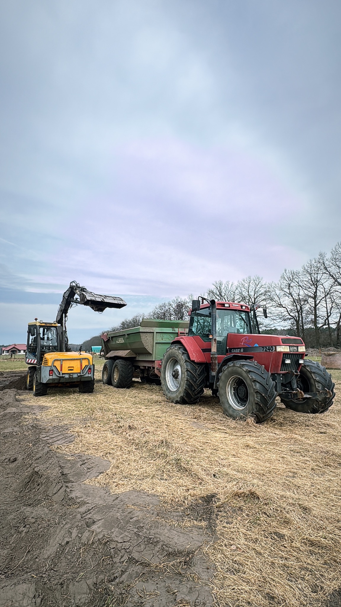 Żółta koparko-ładowarka i czerwony traktor z przyczepą na polu. Koparka nabiera ziemię, sugerując prace ziemne lub przygotowanie terenu pod uprawy.