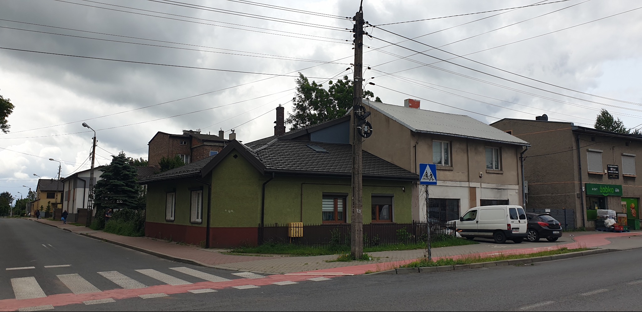 Ujęcie z poziomu ulicy na dom z ciemną dachówką i zieloną elewacją. Widoczne fragmenty innych budynków i słup elektryczny. Pochmurne niebo w tle.