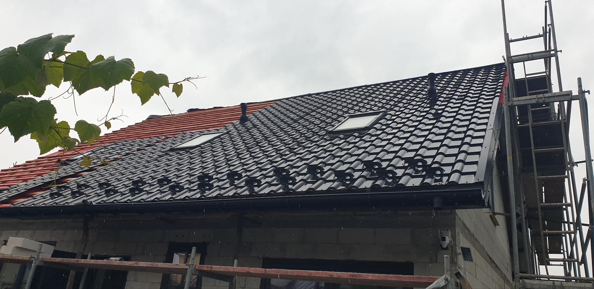 Częściowo pokryty dach z czarną dachówką z oknami dachowymi, obok fragment dachu z czerwoną dachówką, rusztowanie z boku budynku w pochmurny dzień.