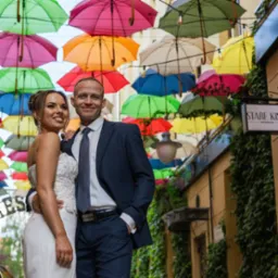 Para młoda w eleganckich strojach pozuje pod kolorowymi parasolkami, dekoracja uliczna. Uśmiechnięci, w tle budynki z zielenią. Romantyczna atmosfera, ciepłe światło.