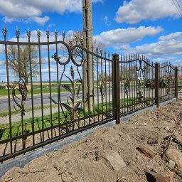 Usługi Spawalnicze Krzysztof Skorniak - Kute ogrodzenie z motywem roślinnym, złocone zdobienia na tle błękitnego nieba. Widoczne słupy energetyczne i fragment drogi. Montaż w toku, obok ziemia i gruz.