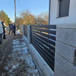 Usługi Spawalnicze Krzysztof Skorniak - Nowoczesne ogrodzenie z poziomych, grafitowych paneli między kamiennymi słupkami, z fragmentem ośnieżonego chodnika i sylwetkami osób w tle.