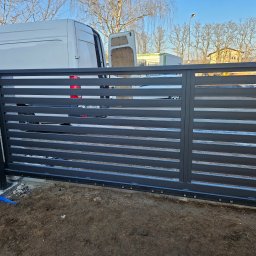Usługi Spawalnicze Krzysztof Skorniak - Nowoczesna, przesuwna brama wjazdowa z poziomymi panelami w kolorze grafitowym, widoczna na tle białego samochodu dostawczego i drzew bez liści.