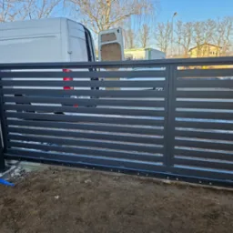 Nowoczesna, przesuwna brama wjazdowa z poziomymi panelami w kolorze grafitowym, widoczna na tle białego samochodu dostawczego i drzew bez liści.