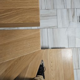 Usługi Spawalnicze Krzysztof Skorniak - Drewniane schody z widocznym fragmentem podłogi wyłożonej płytkami imitującymi marmur. U dołu kadru widoczne stopy osoby stojącej na schodach.