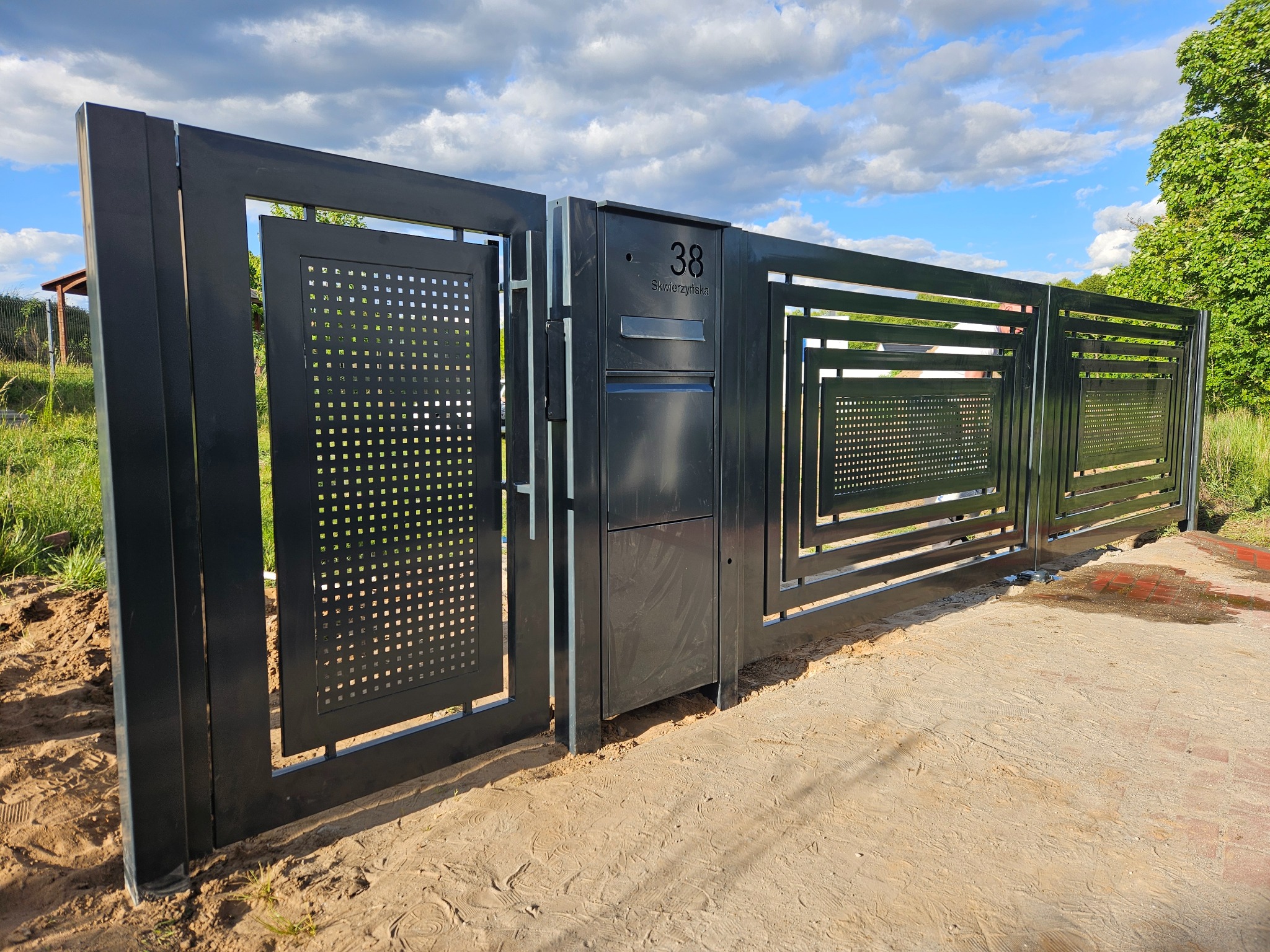 Nowoczesna, grafitowa brama wjazdowa z perforowanymi panelami i numerem domu '38 Skwierzyńska', na tle błękitnego nieba z chmurami.