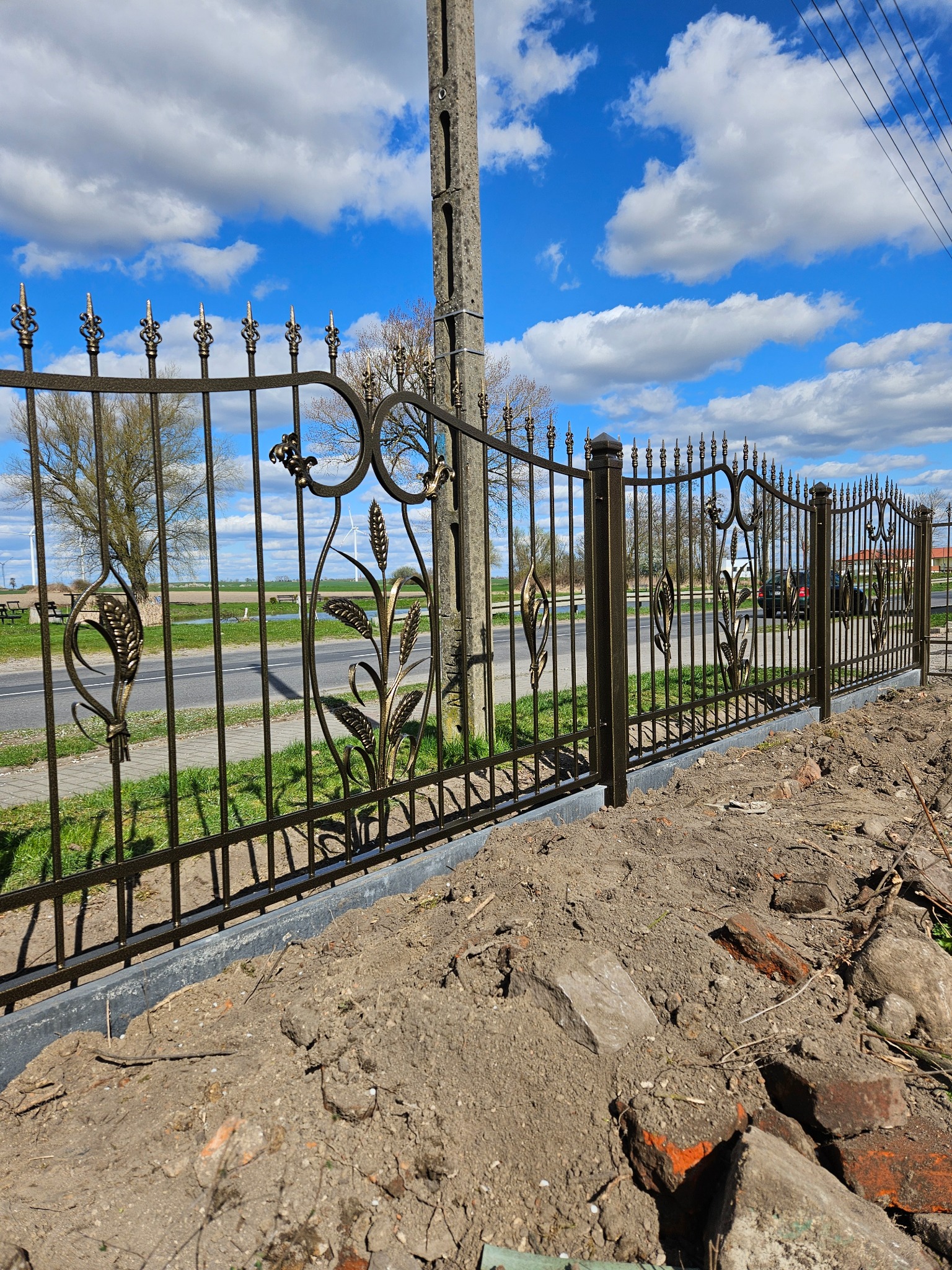 Kute ogrodzenie z motywem roślinnym, złocone zdobienia na tle błękitnego nieba. Widoczne słupy energetyczne i fragment drogi. Montaż w toku, obok ziemia i gruz.