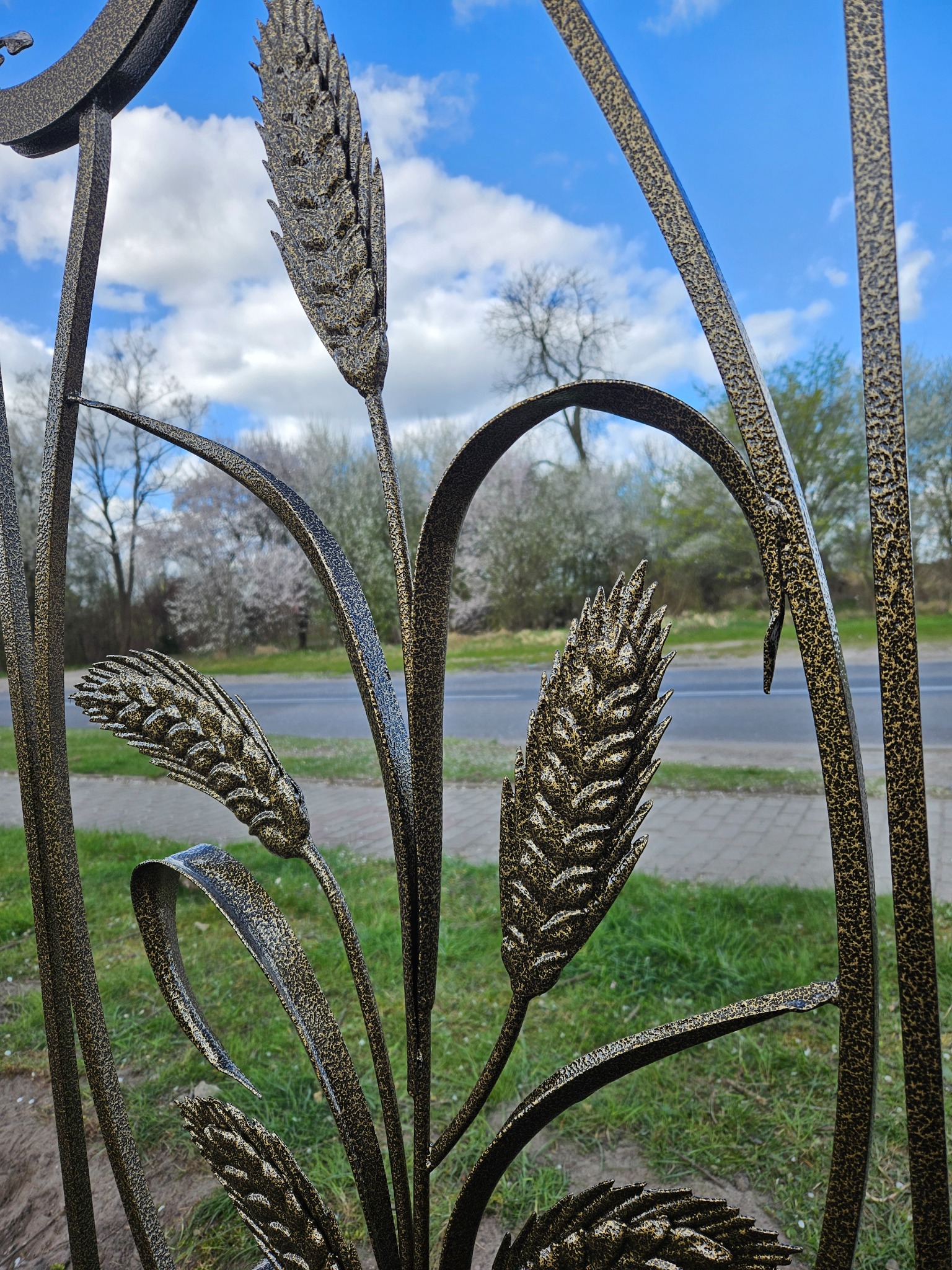 Kute ogrodzenie z motywem pszenicy, złoto-czarna kolorystyka, na tle błękitnego nieba z chmurami i zieleni. Detal artystyczny.