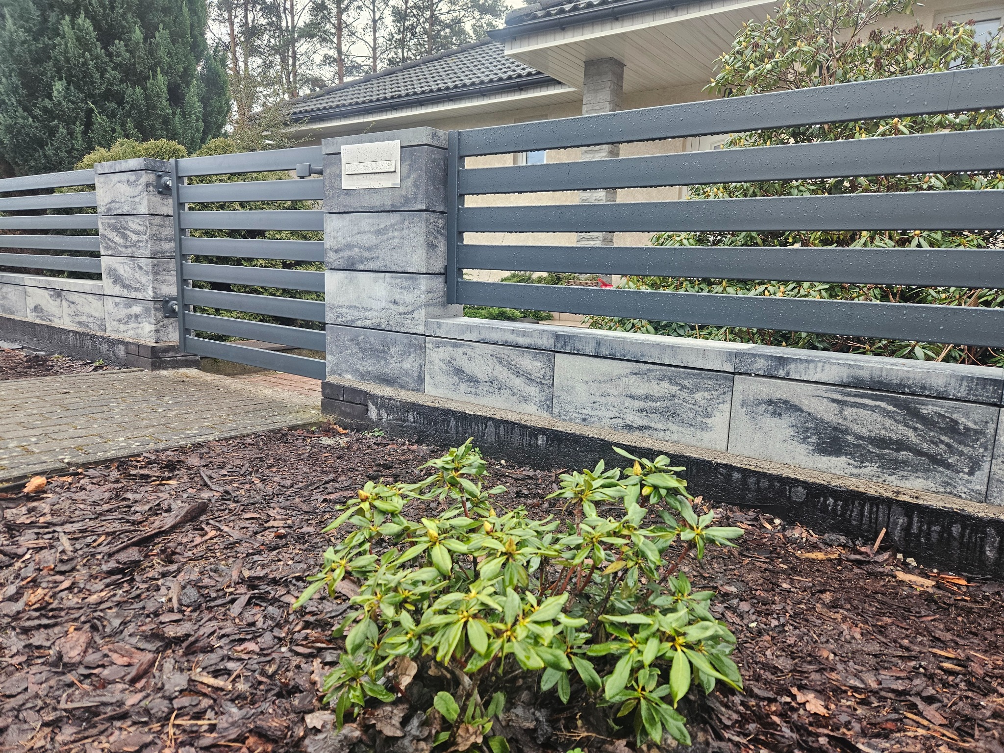 Nowoczesne ogrodzenie z szarych, poziomych paneli i kamiennych słupków. Widoczne krople deszczu. W tle dom i roślinność. Ujęcie z poziomu gruntu.