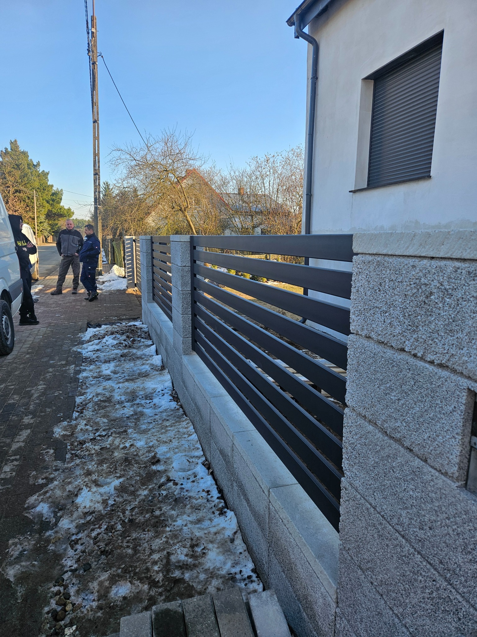 Nowoczesne ogrodzenie z poziomych, grafitowych paneli między kamiennymi słupkami, z fragmentem ośnieżonego chodnika i sylwetkami osób w tle.