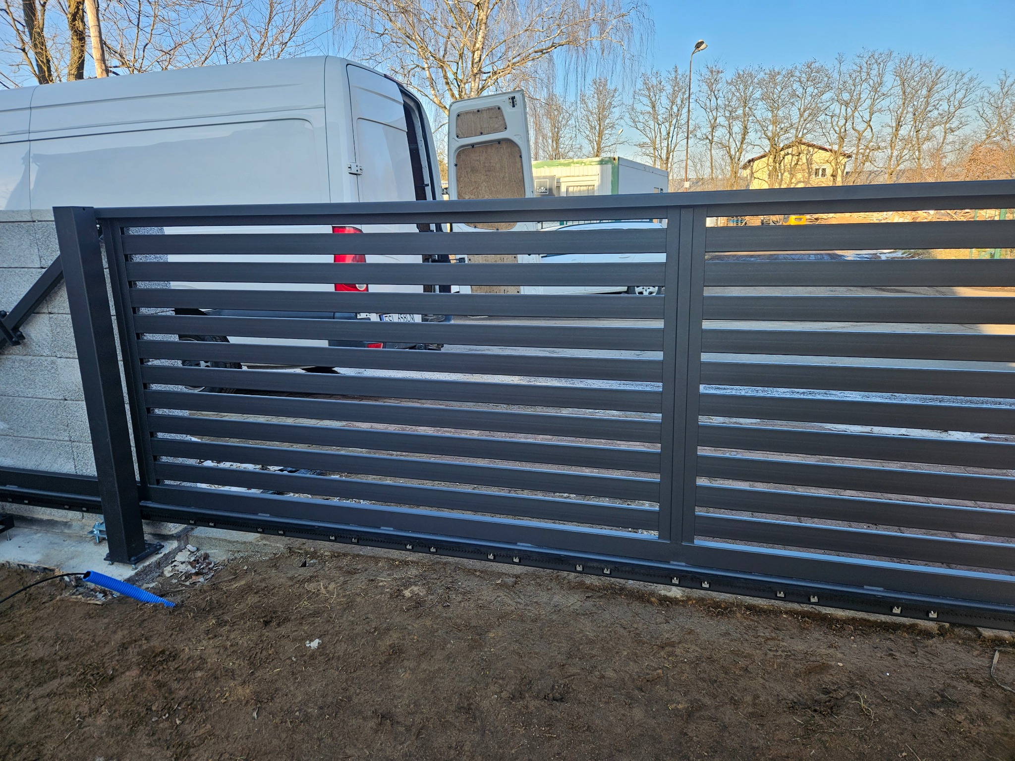 Nowoczesna, przesuwna brama wjazdowa z poziomymi panelami w kolorze grafitowym, widoczna na tle białego samochodu dostawczego i drzew bez liści.