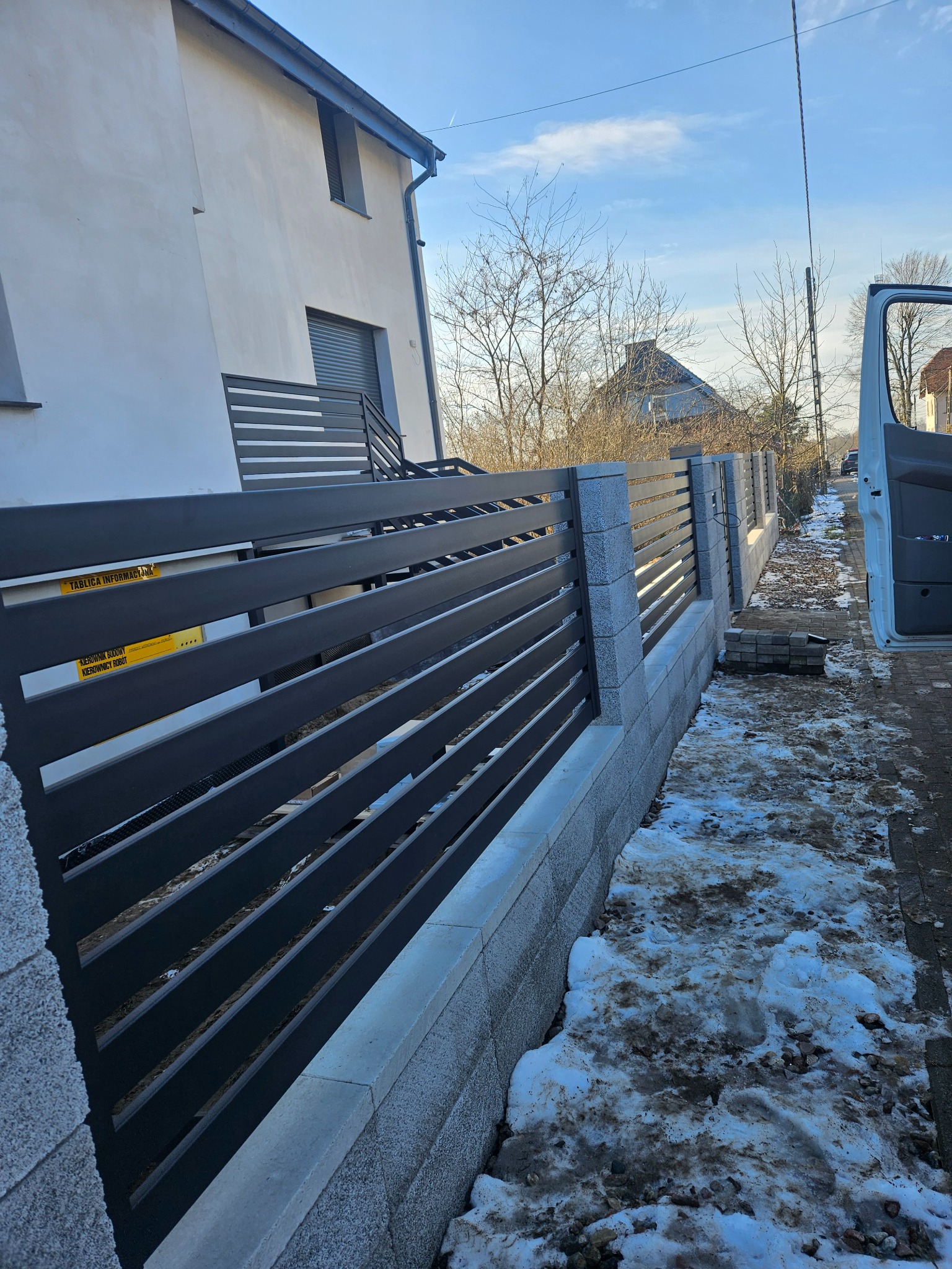 Nowoczesne ogrodzenie posesji z poziomych, grafitowych paneli i szarych słupków z betonu architektonicznego, z widoczną tablicą informacyjną budowy.