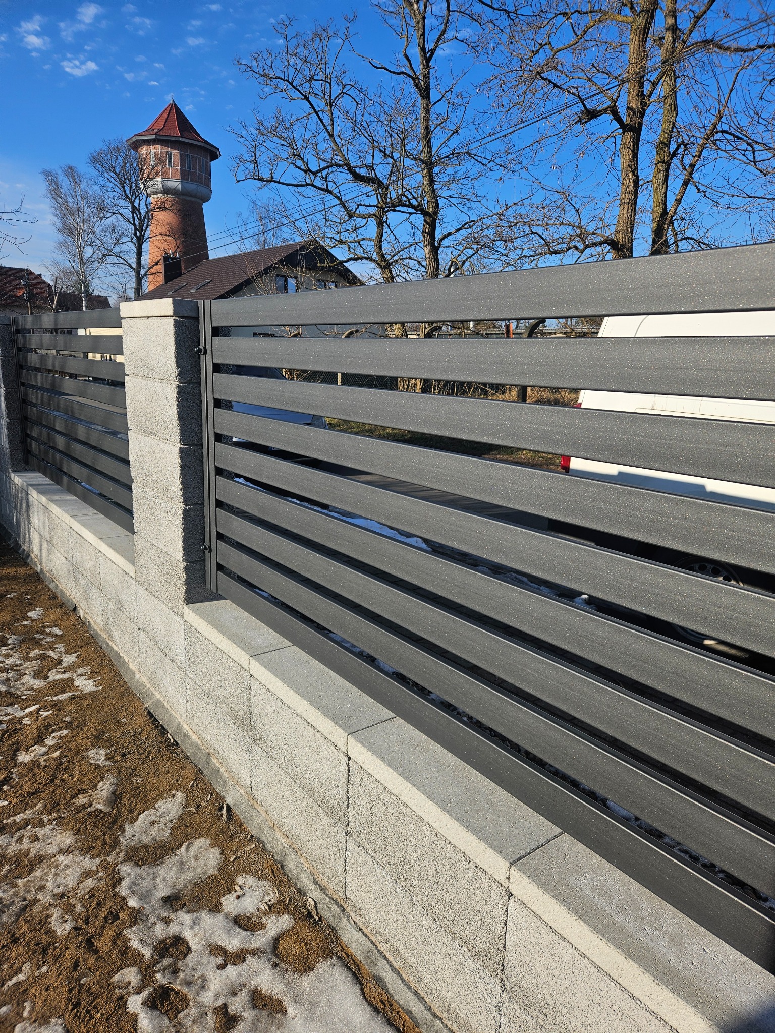 Nowoczesne ogrodzenie panelowe z szarego kompozytu na betonowej podmurówce, widok z boku, w tle wieża ciśnień i błękitne niebo.