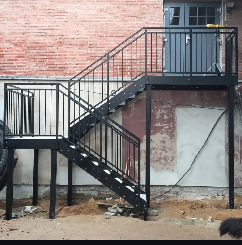 Zewnętrzne, czarne schody metalowe z poręczą prowadzące do drzwi na podwyższeniu, z ceglanym murem w tle. Widoczne ślady prac ziemnych i fragment czarnego węża karbowanego.
