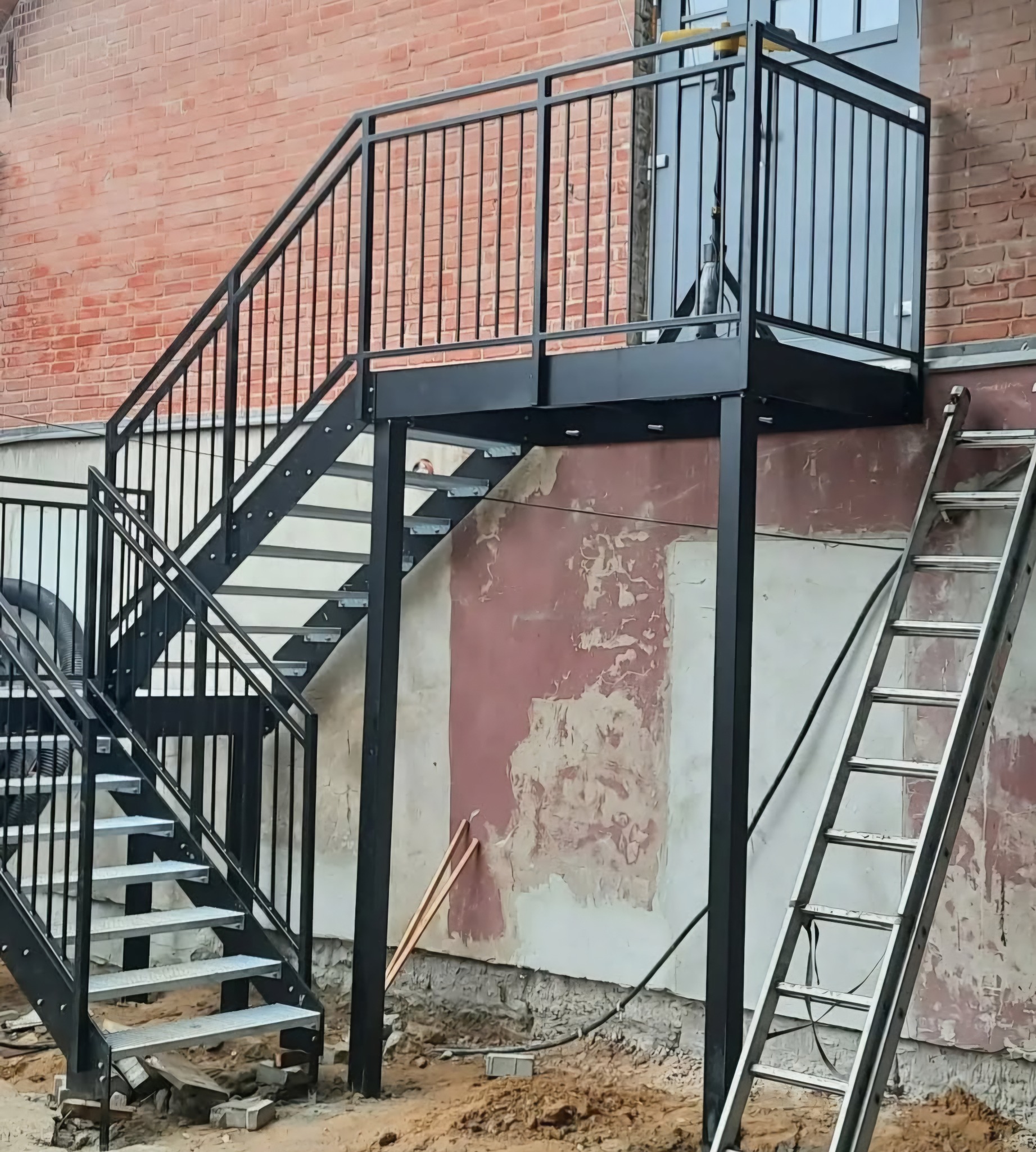 Zewnętrzne schody metalowe z czarną balustradą, prowadzące do drzwi na podwyższeniu. Obok oparta drabina, ściana z cegły i łuszcząca się farba. Widoczne narzędzia.