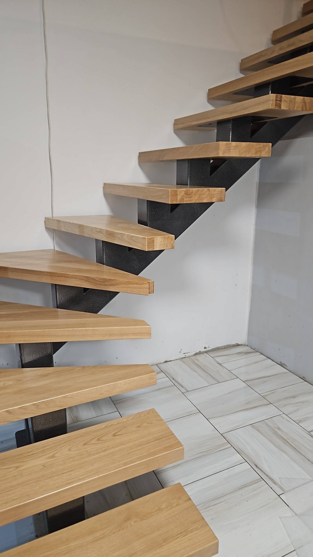 Nowoczesne schody z drewnianymi stopniami i czarną, metalową konstrukcją, widok od dołu, ukazujący detale wykończenia i połączenia elementów.
