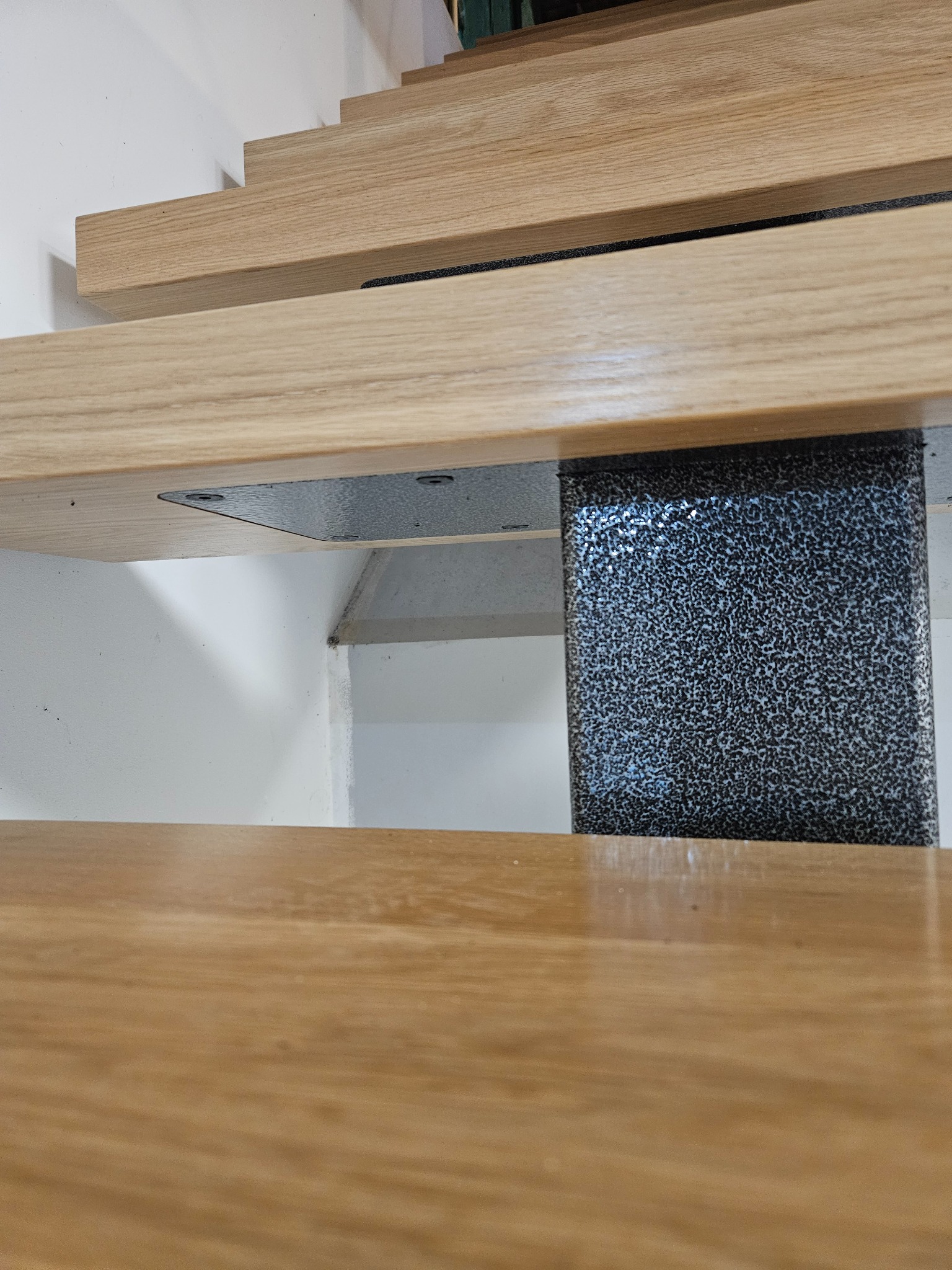 Detale drewnianych schodów z metalowym wspornikiem o teksturowanej powierzchni, widoczne mocowania śrubowe, jasne oświetlenie, minimalistyczny design.