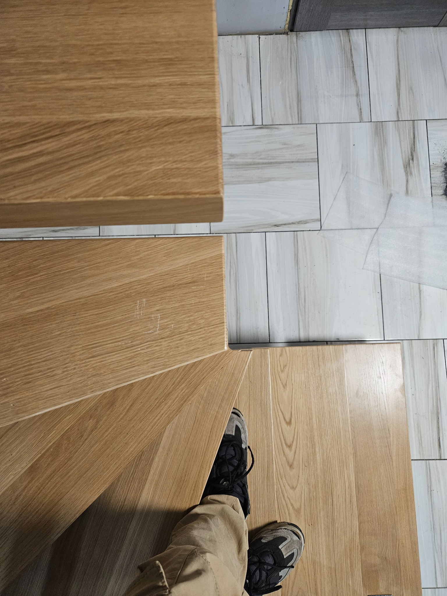 Drewniane schody z widocznym fragmentem podłogi wyłożonej płytkami imitującymi marmur. U dołu kadru widoczne stopy osoby stojącej na schodach.