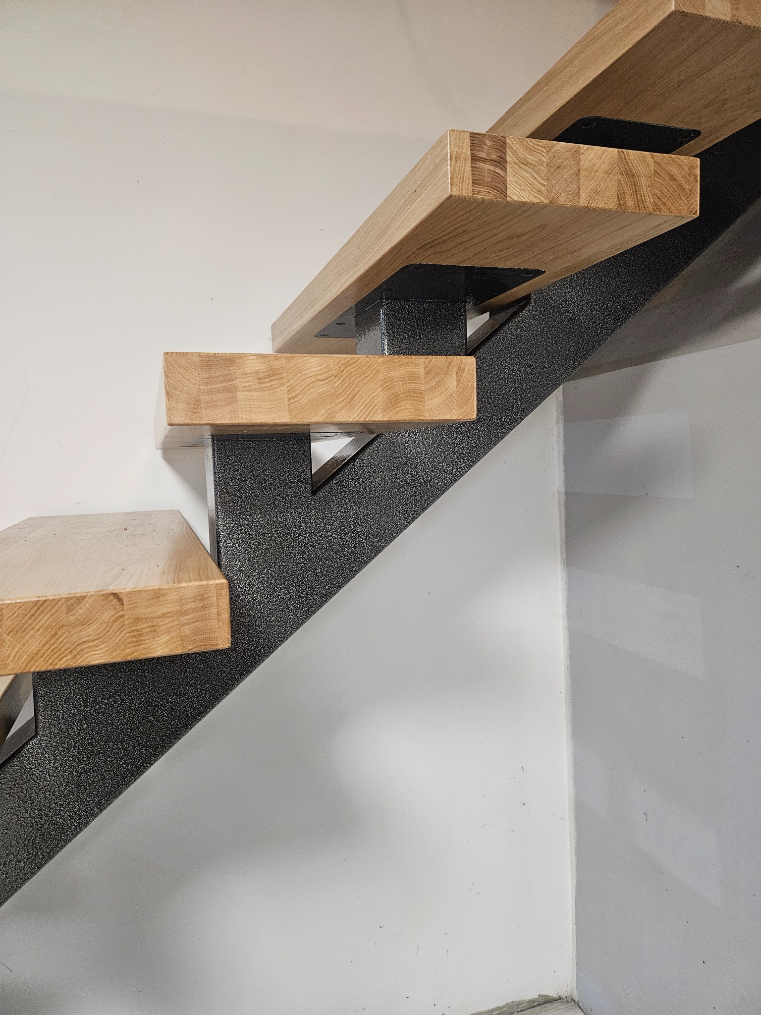 Nowoczesne schody z drewnianymi stopniami i metalową konstrukcją o fakturze piasku, umieszczone w rogu pomieszczenia z białymi ścianami.