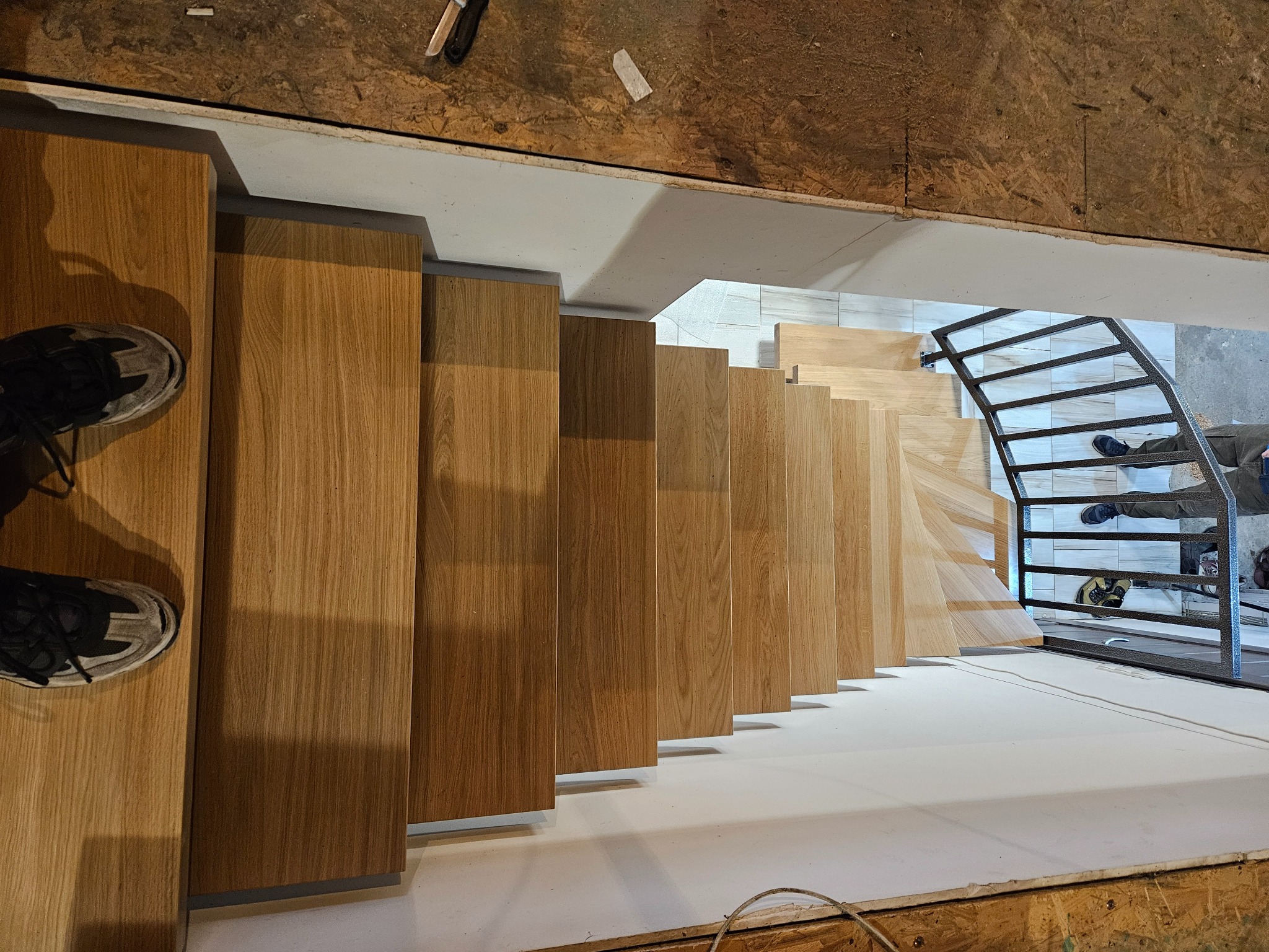 Nowoczesne schody drewniane z metalową balustradą w trakcie montażu. Widok z góry, ukazujący detale wykończenia stopni i konstrukcji, z butami na pierwszym planie.