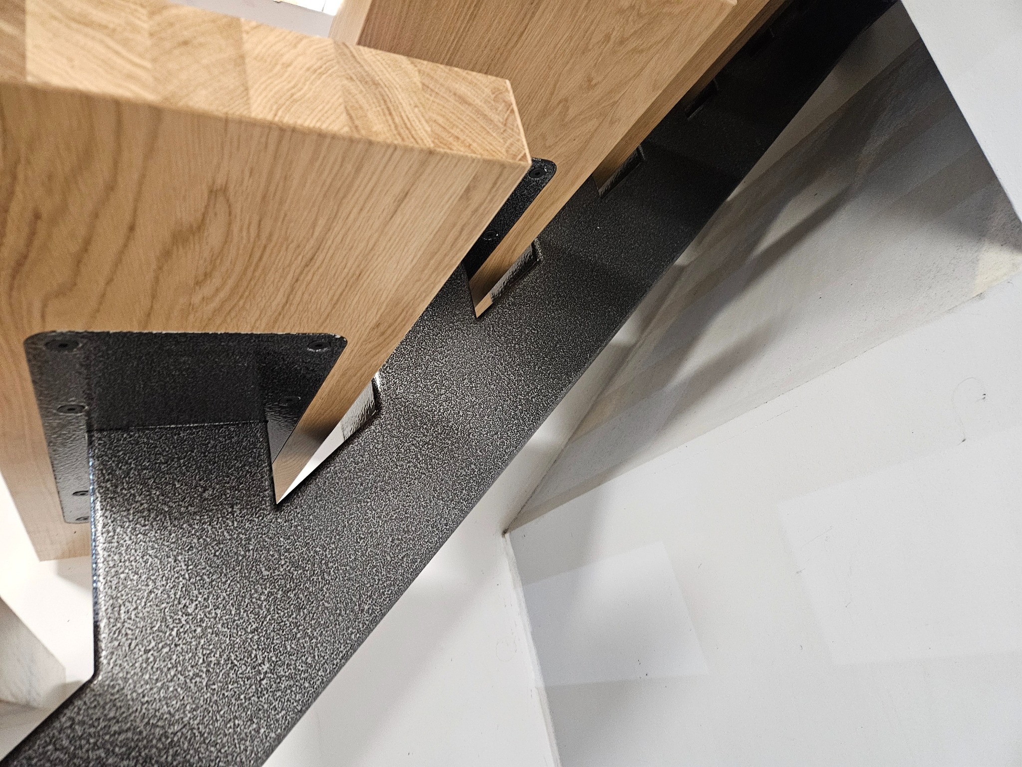 Detal drewnianych schodów z metalową konstrukcją nośną. Widoczne łączenie stopnia z elementem wspornym. Minimalistyczny design, jasne drewno i grafitowa stal.