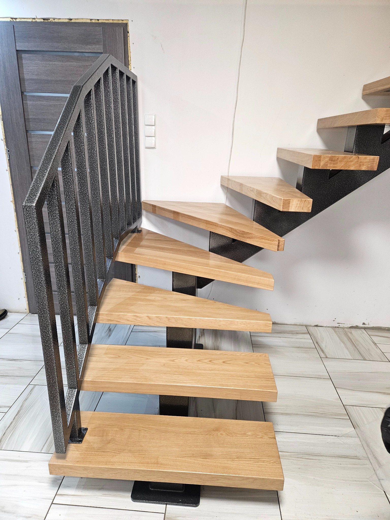 Nowoczesne schody z metalową poręczą i drewnianymi stopniami, z boku widoczne drzwi. Minimalistyczny design, jasne wnętrze z płytkami podłogowymi.