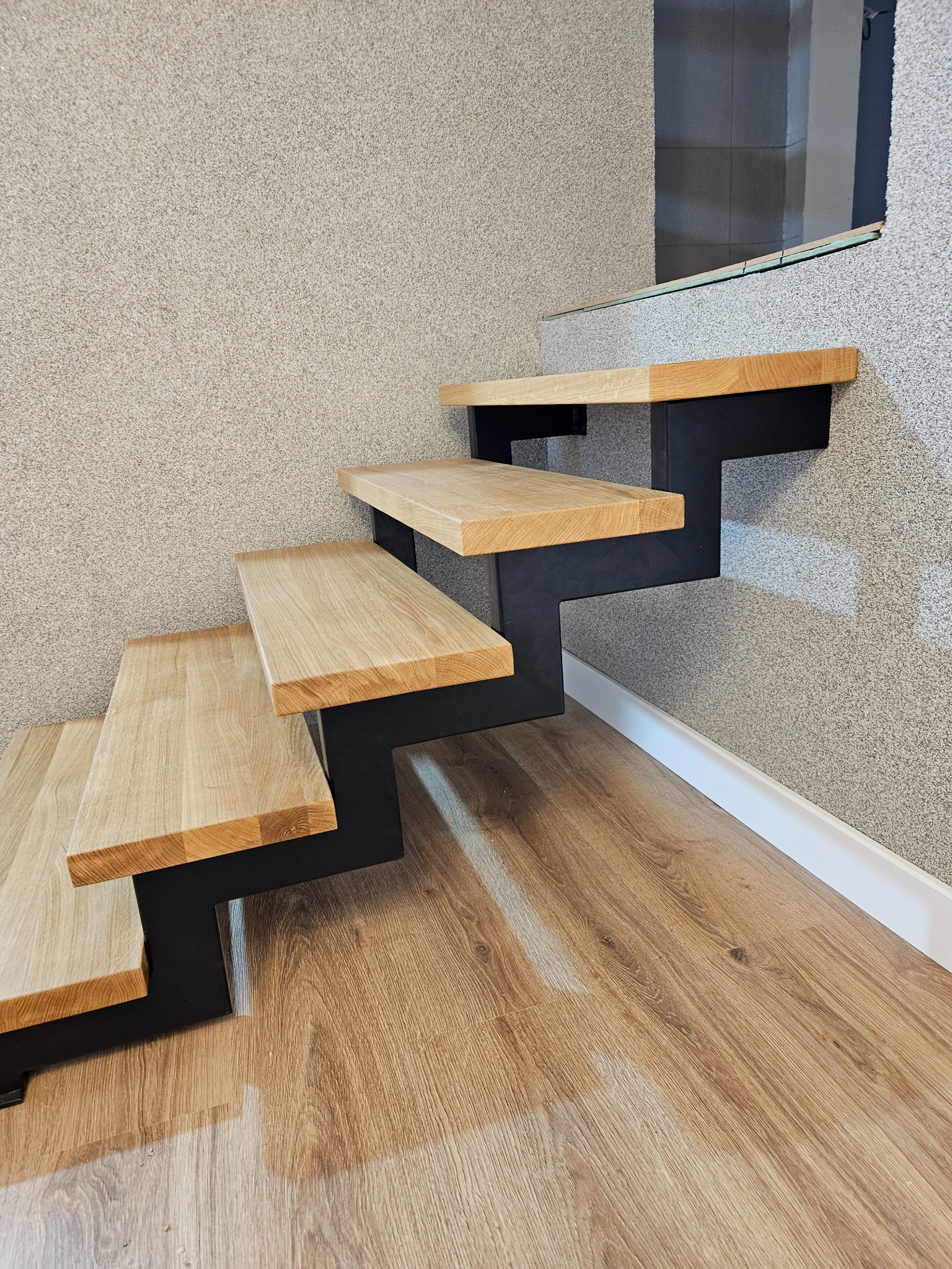 Nowoczesne schody: drewniane stopnie na czarnych, metalowych wspornikach. Minimalistyczny design, jasne drewno, szara ściana i podłoga z paneli.