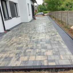 Ułożona kostka brukowa w odcieniach szarości i beżu tworzy wzór na podjeździe. W tle budynek z białymi ścianami i betonowe ogrodzenie. Widoczny etap prac wykończeniowych.