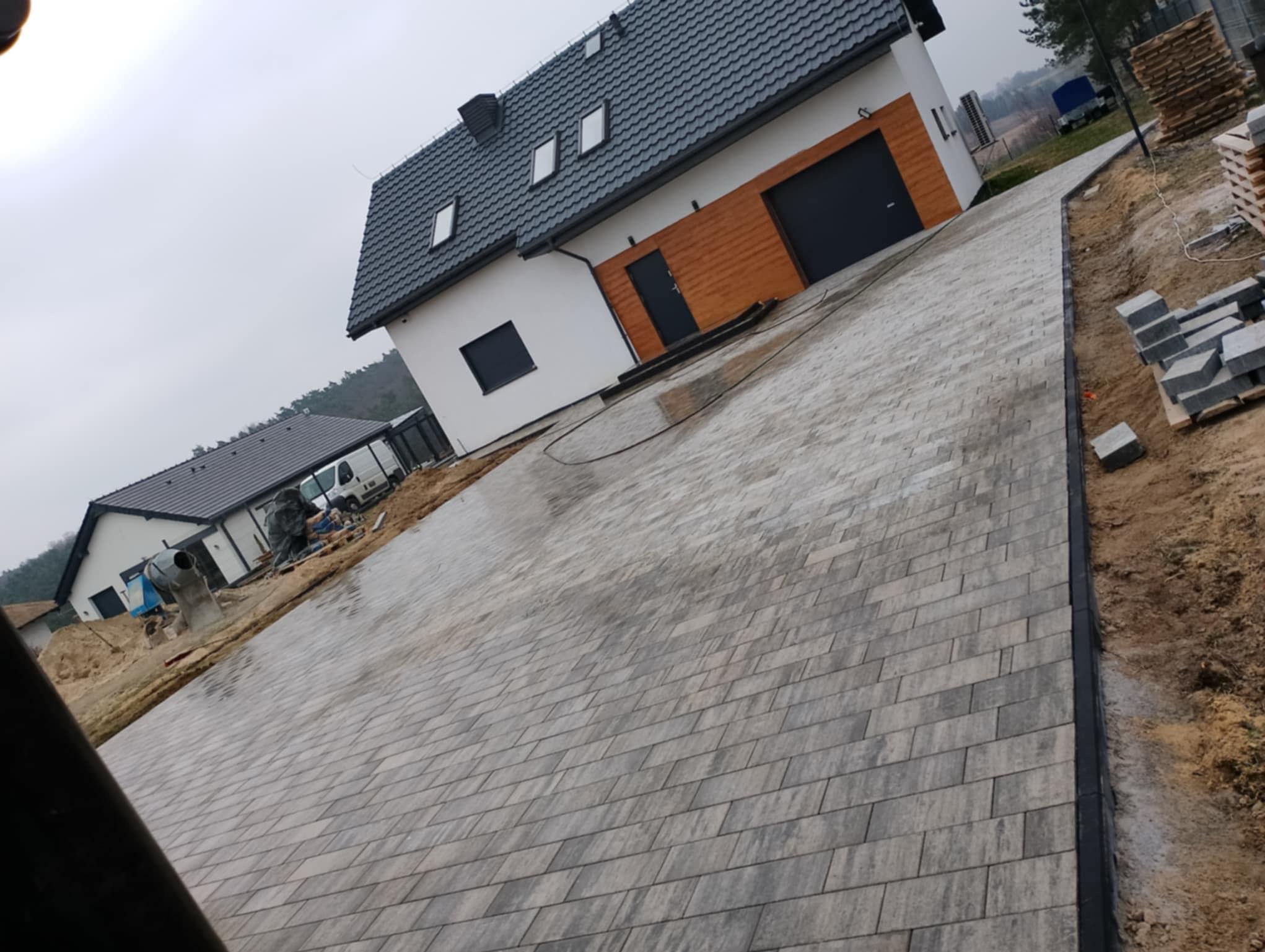 Ułożona szara kostka brukowa przed nowoczesnym domem z garażem, widoczne krawężniki i przygotowane materiały. Ujęcie z niskiego kąta.