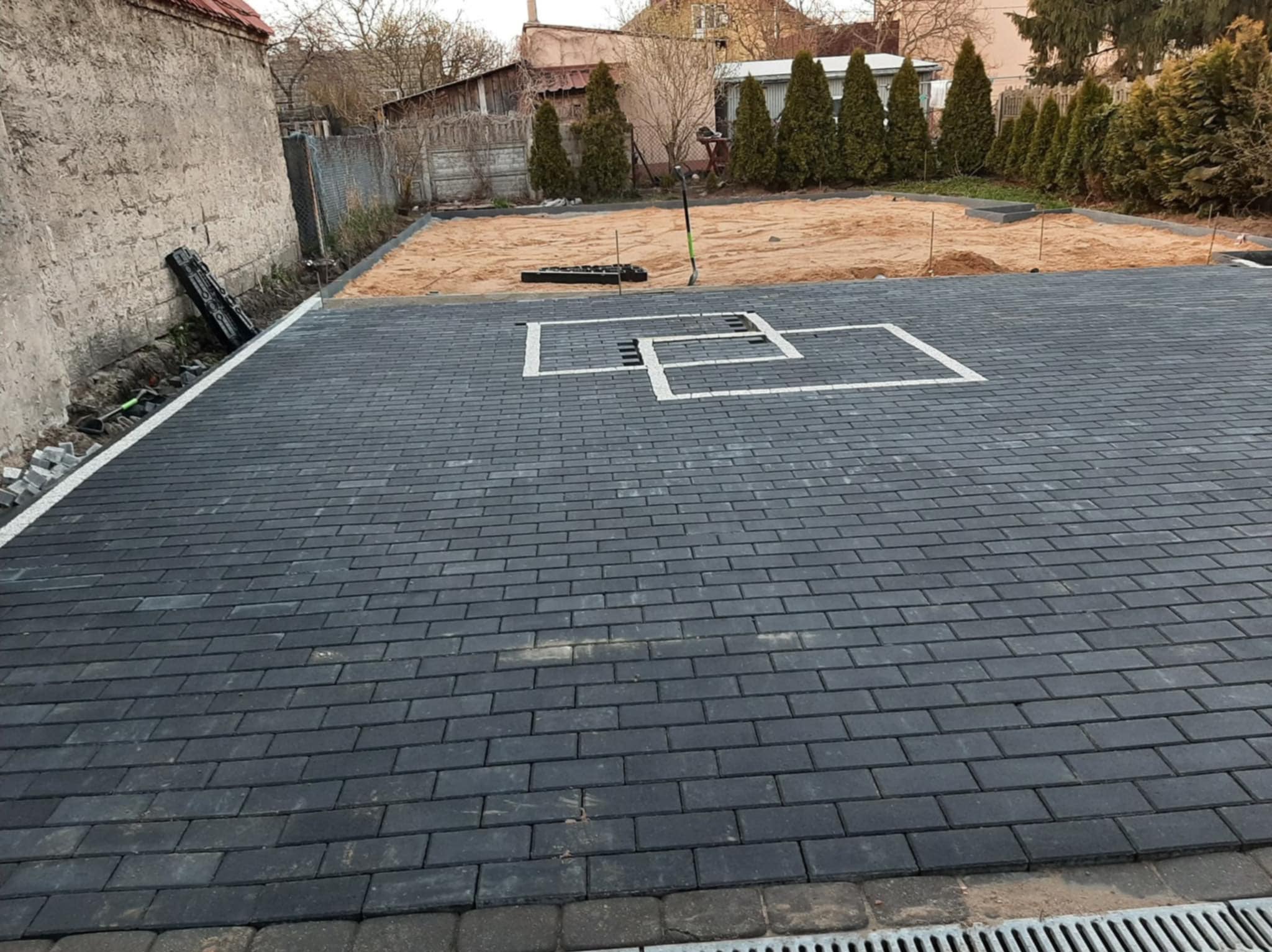 Ułożona, grafitowa kostka brukowa z białymi elementami dekoracyjnymi na podjeździe. W tle teren przygotowany pod dalsze prace brukarskie.