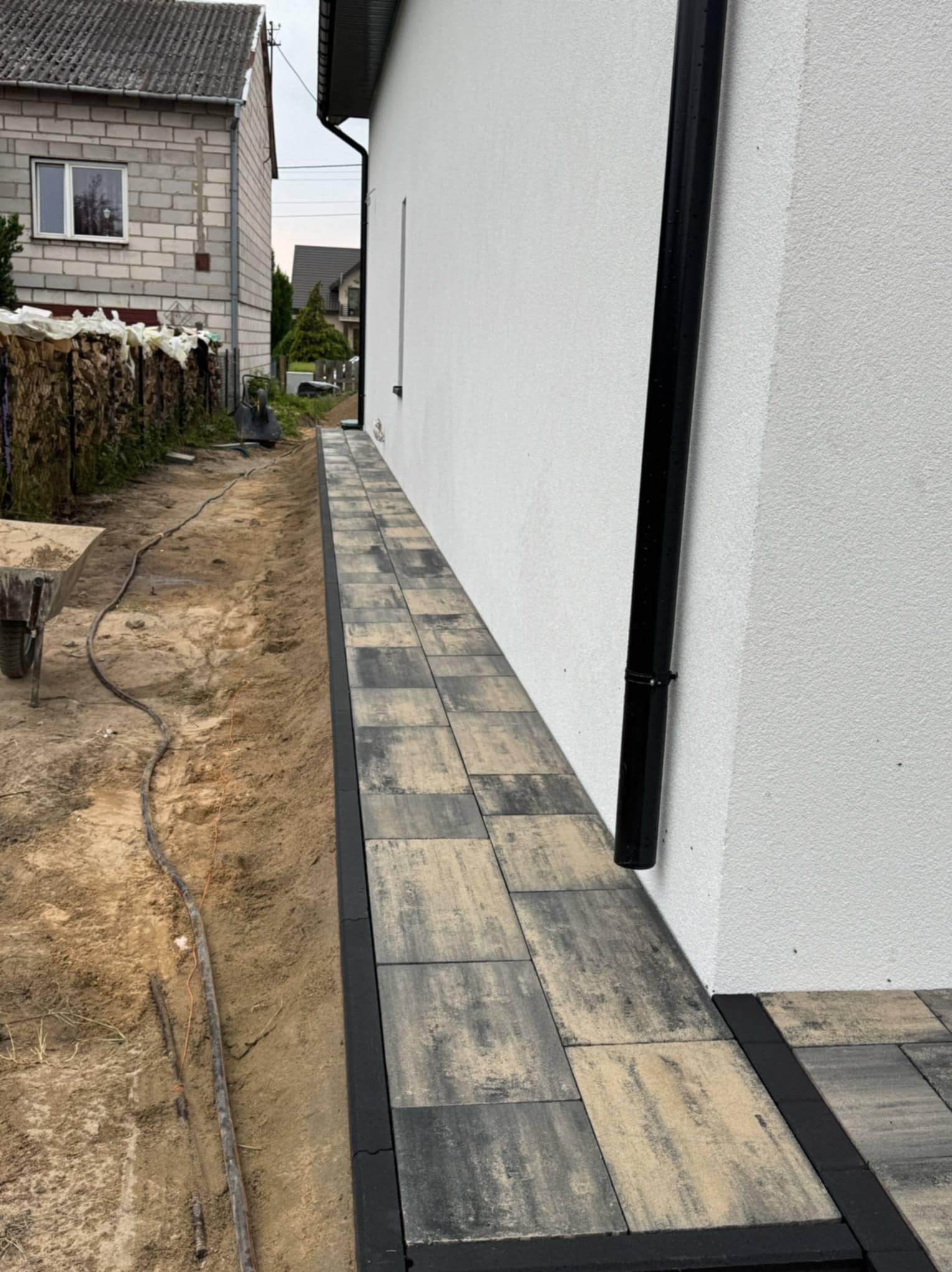 Ułożona kostka brukowa wzdłuż białej ściany domu z czarną rynną. Widoczny fragment posesji w trakcie prac. Kostka w odcieniach szarości i beżu.