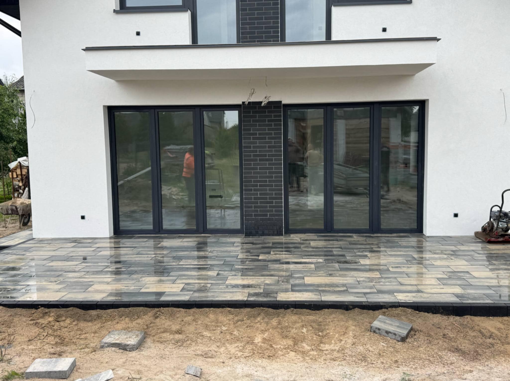 Nowoczesny taras z kostki brukowej w odcieniach szarości, przylegający do białego budynku z dużymi, przesuwnymi drzwiami. Częściowo wykończony teren wokół tarasu.