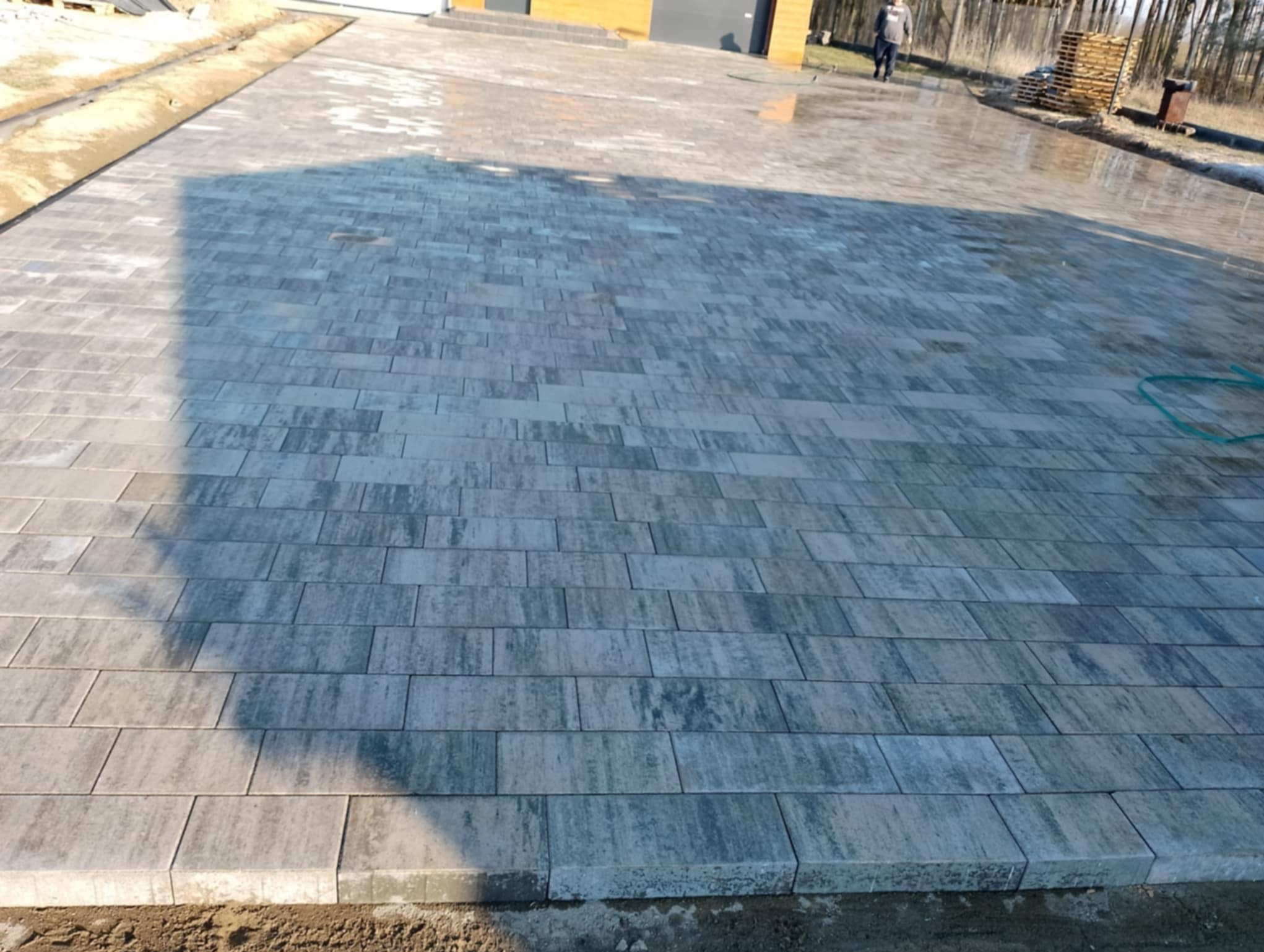Ułożona szara kostka brukowa na posesji, widoczne obrzeża i przygotowane podłoże. W tle budynek z garażem i osoba. Całość w słoneczny dzień.