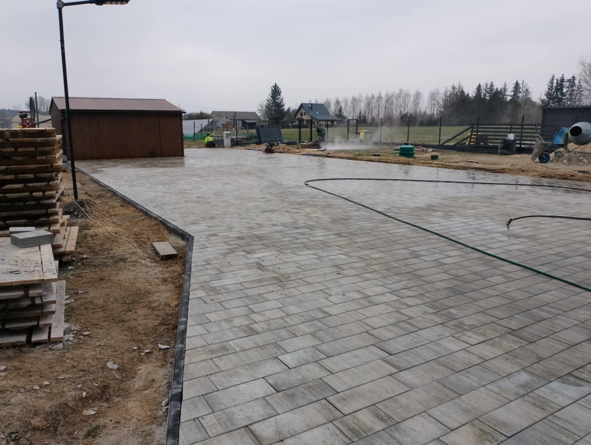 Ułożona, szara kostka brukowa na podjeździe, widoczny fragment ogrodu z elementami ogrodzenia i budynkami w tle. Prace brukarskie w toku, widoczny wąż do podlewania.