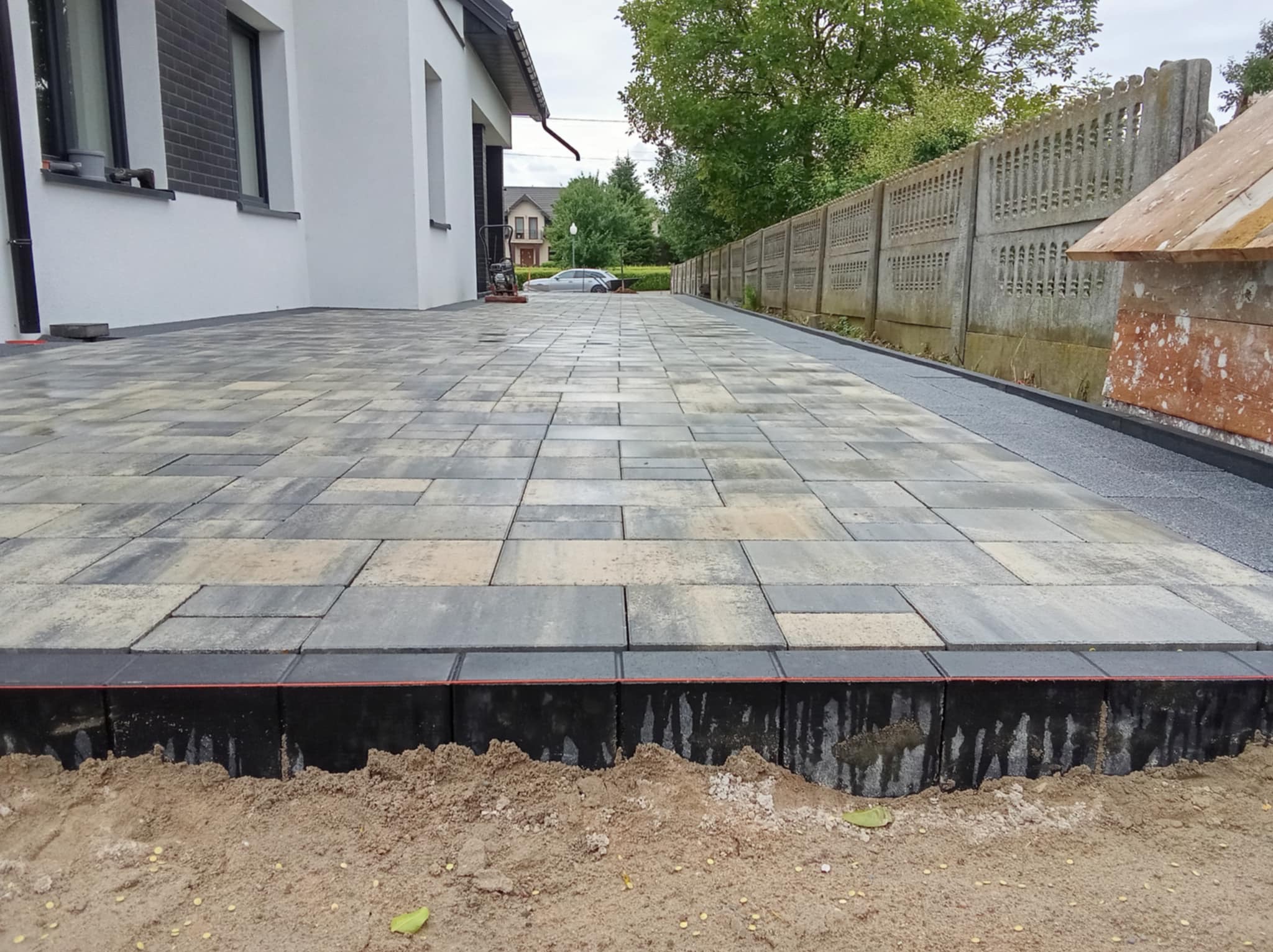 Ułożona, wielokolorowa kostka brukowa na podjeździe domu z szarą obwódką. W tle ogrodzenie z betonowych płyt i budynek mieszkalny.