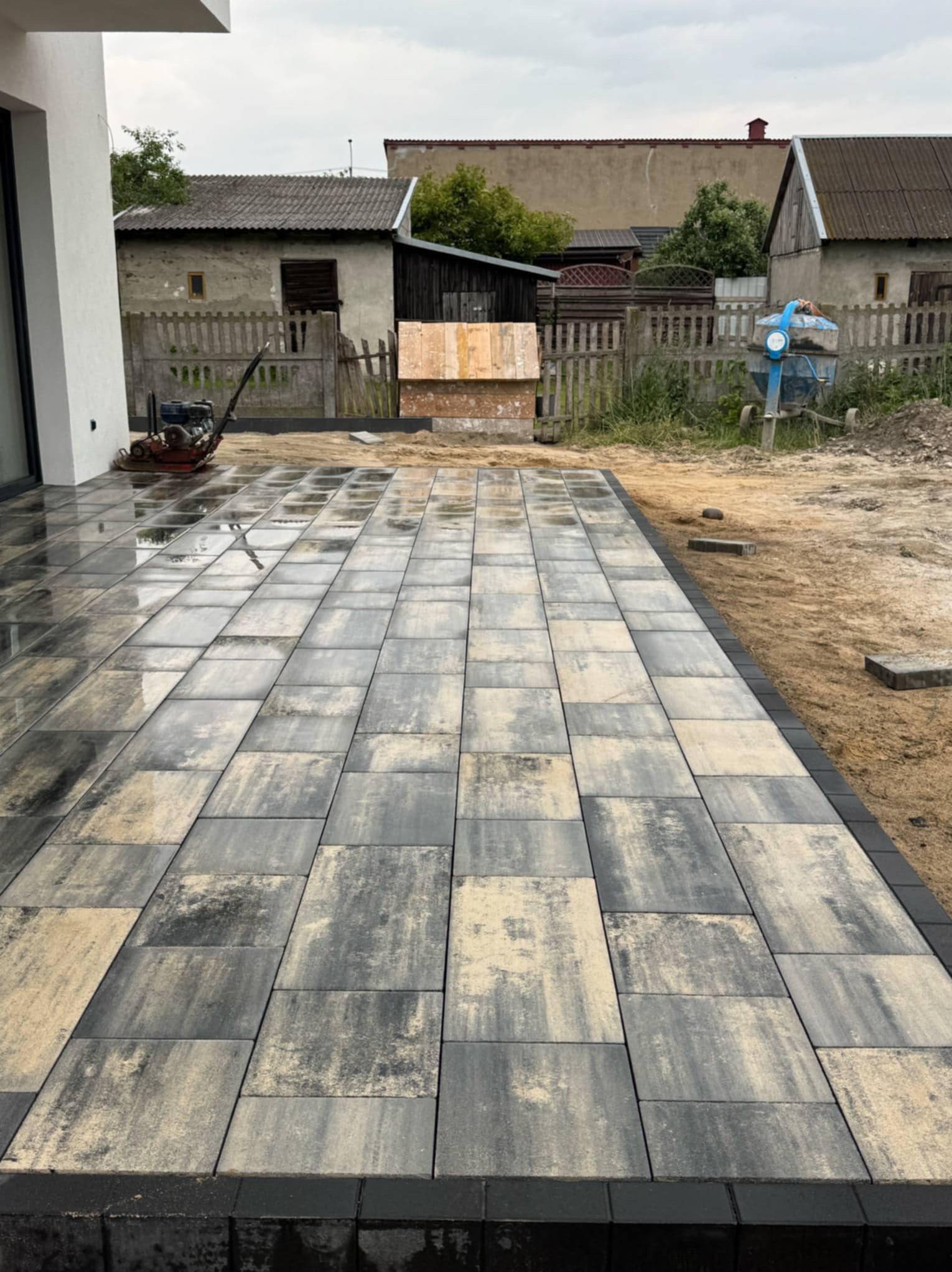 Ułożona, prostokątna kostka brukowa w odcieniach szarości i beżu, z czarnym obrzeżem. W tle budynki gospodarcze i sprzęt budowlany.