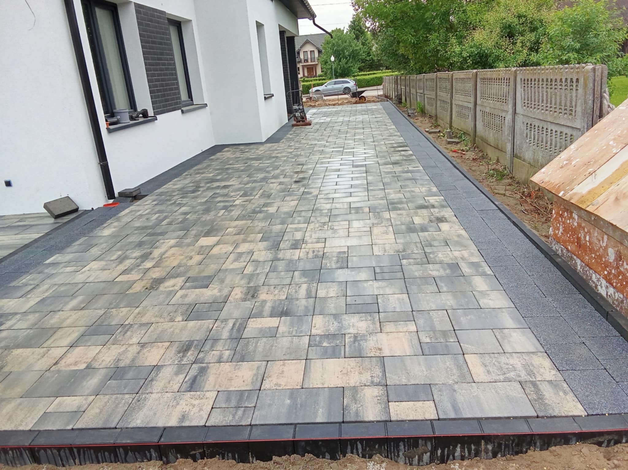 Ułożona kostka brukowa w odcieniach szarości i beżu tworzy wzór na podjeździe. W tle budynek z białymi ścianami i betonowe ogrodzenie. Widoczny etap prac wykończeniowych.