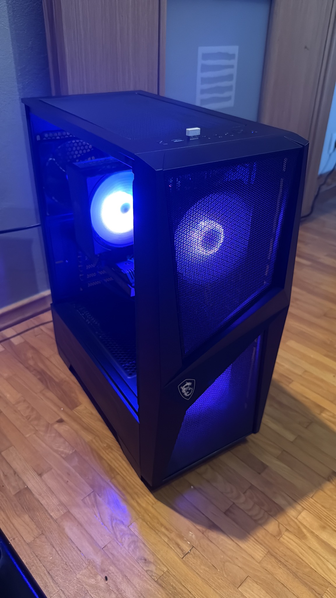 Czarna obudowa komputera PC z podświetleniem LED, widoczne wentylatory i logo MSI, stoi na drewnianej podłodze. W tle ściana i drzwi.