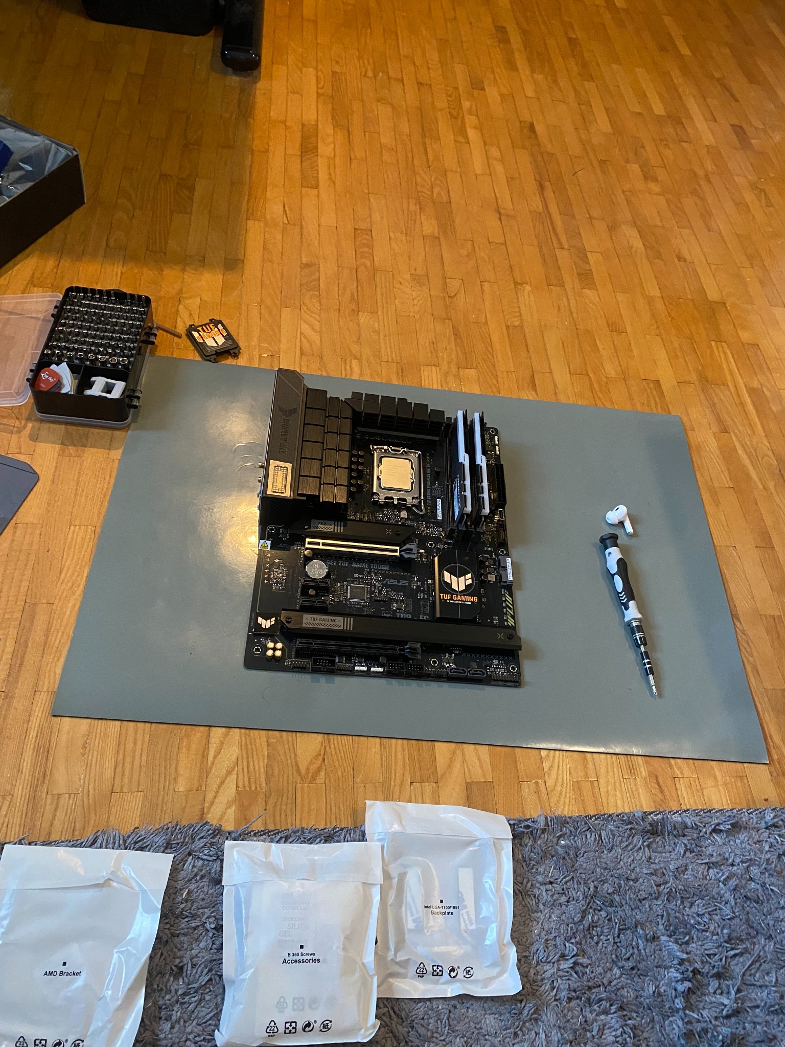 Płyta główna TUF Gaming na macie ESD z akcesoriami, śrubokrętem i słuchawkami, przygotowana do montażu, na drewnianej podłodze. Widoczne opakowania z akcesoriami.
