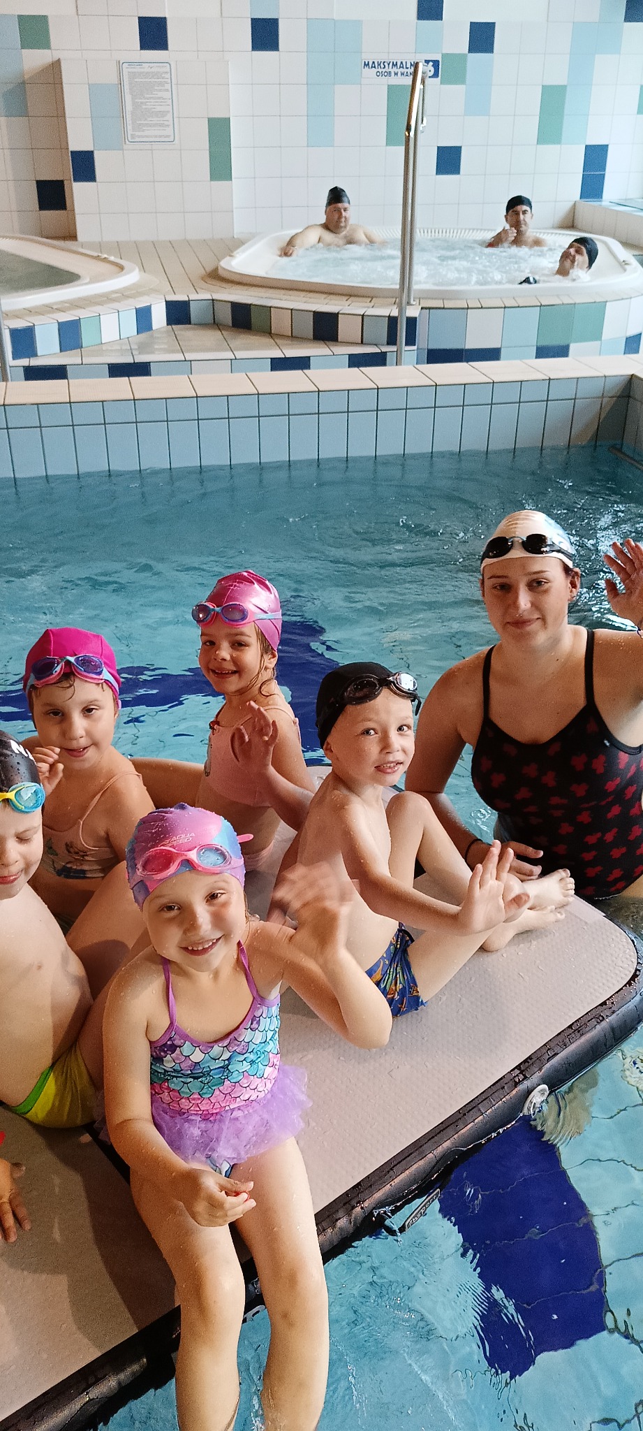 Instruktor z grupą uśmiechniętych dzieci w czepkach i okularkach na platformie w basenie. W tle widać osoby w jacuzzi. Lekcja pływania dla najmłodszych.