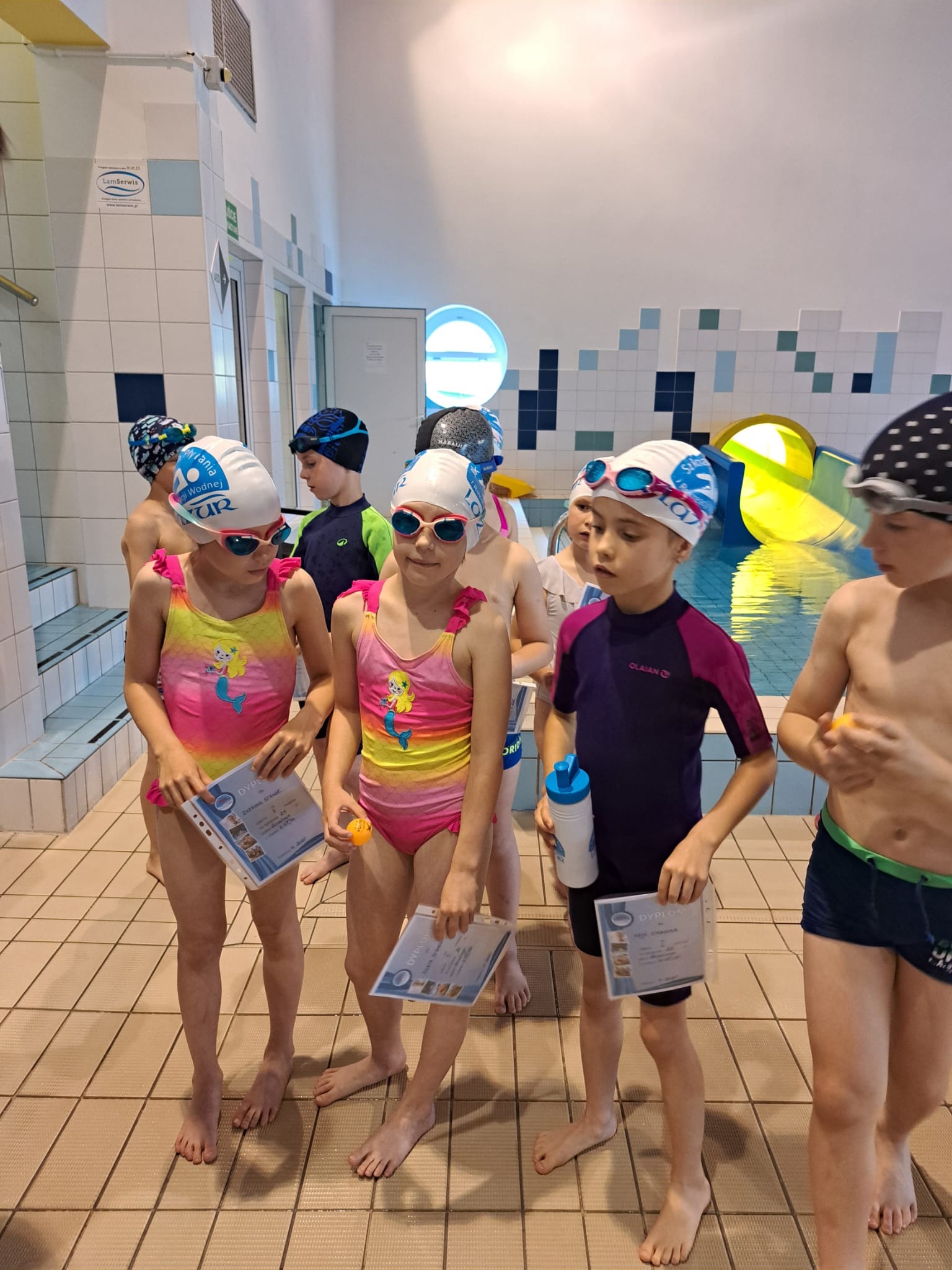 Grupa dzieci w strojach kąpielowych i czepkach, trzymających dyplomy na tle basenu z żółtą zjeżdżalnią. Uczestnicy kursu pływania z nagrodami.