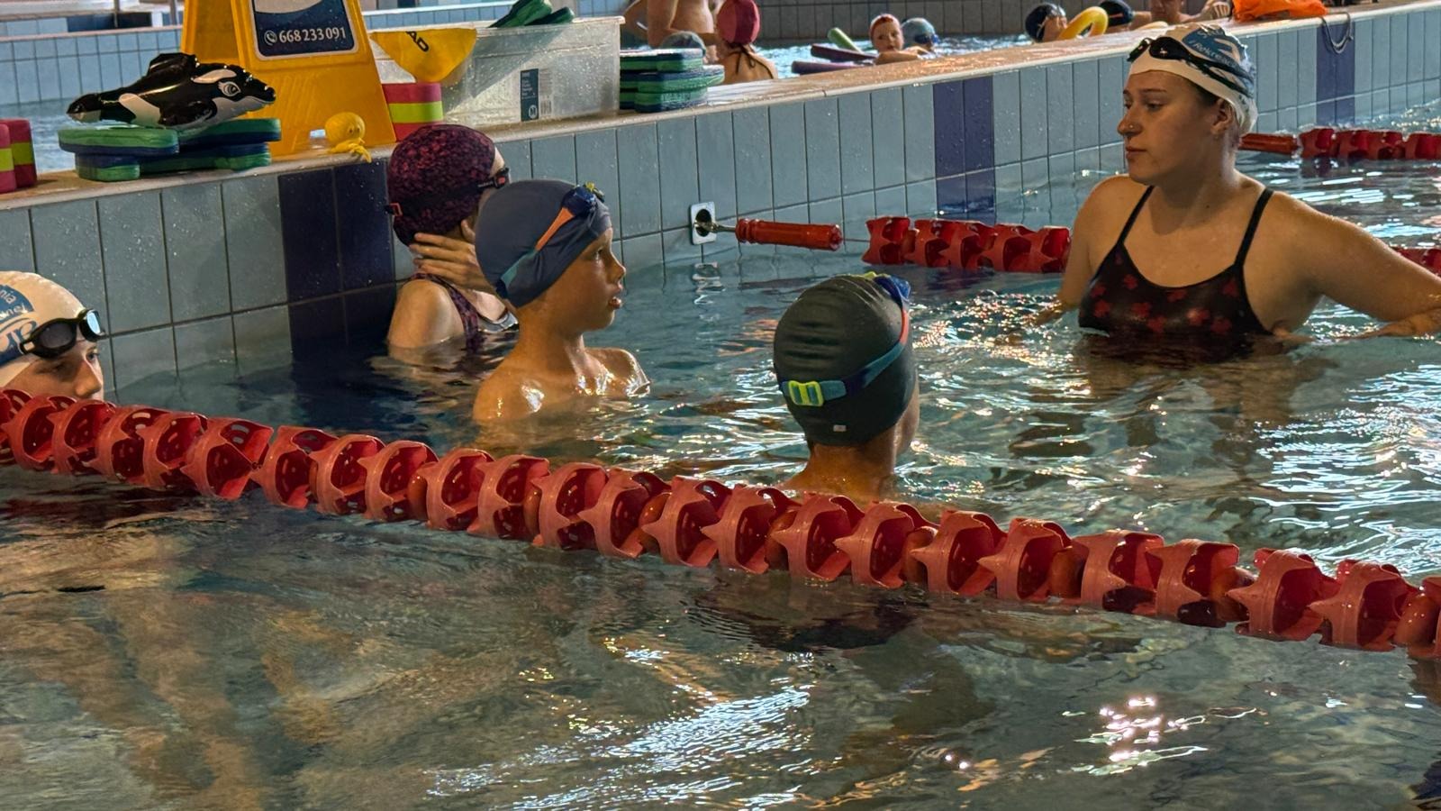 Instruktorka w wodzie z grupą dzieci podczas zajęć na basenie. W tle widoczne akcesoria pływackie i płytki basenowe.